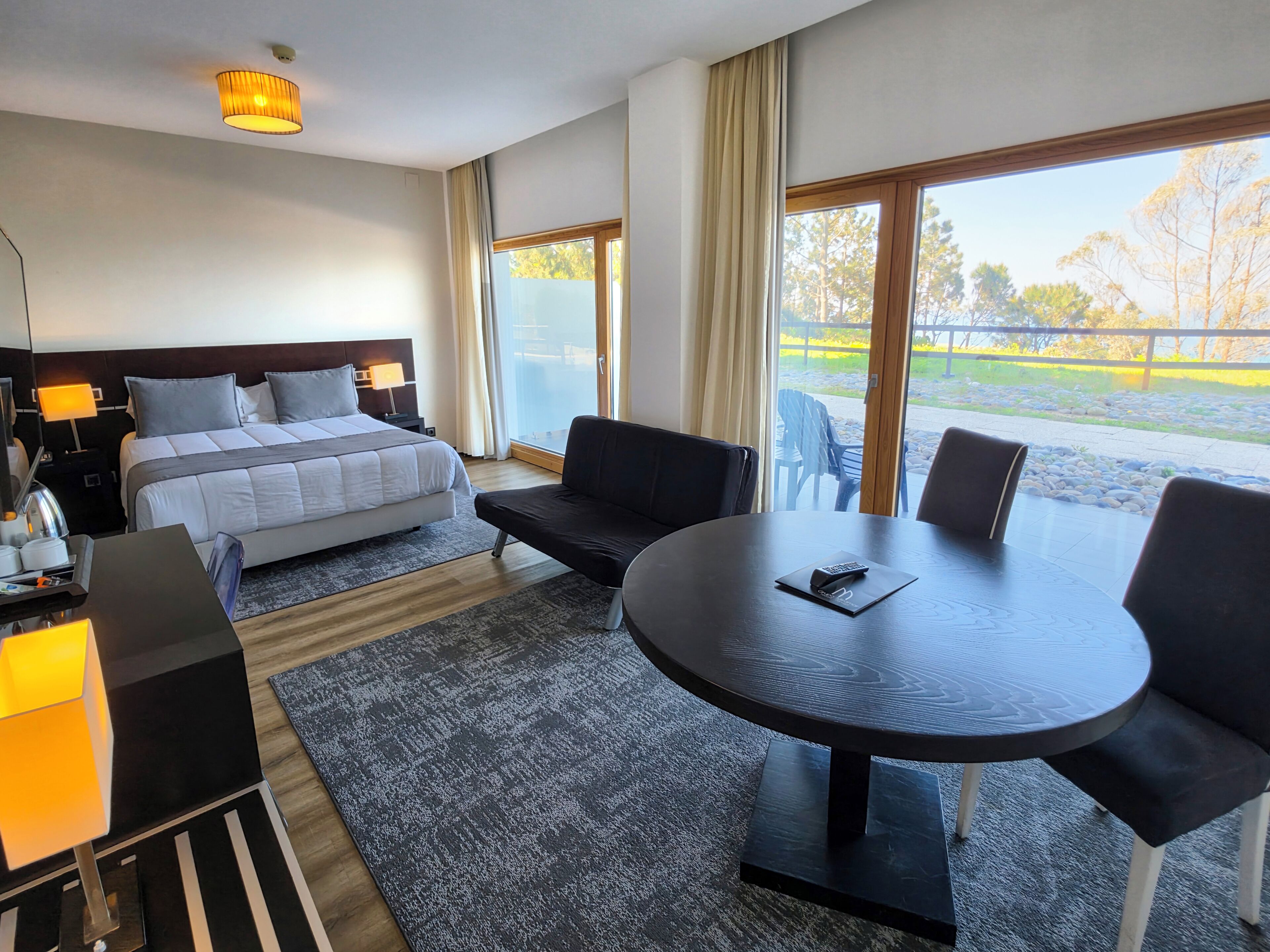 Une chambre d’hôtel avec un lit, un bureau avec une lampe, une table ronde avec des chaises et une vue sur l’extérieur.