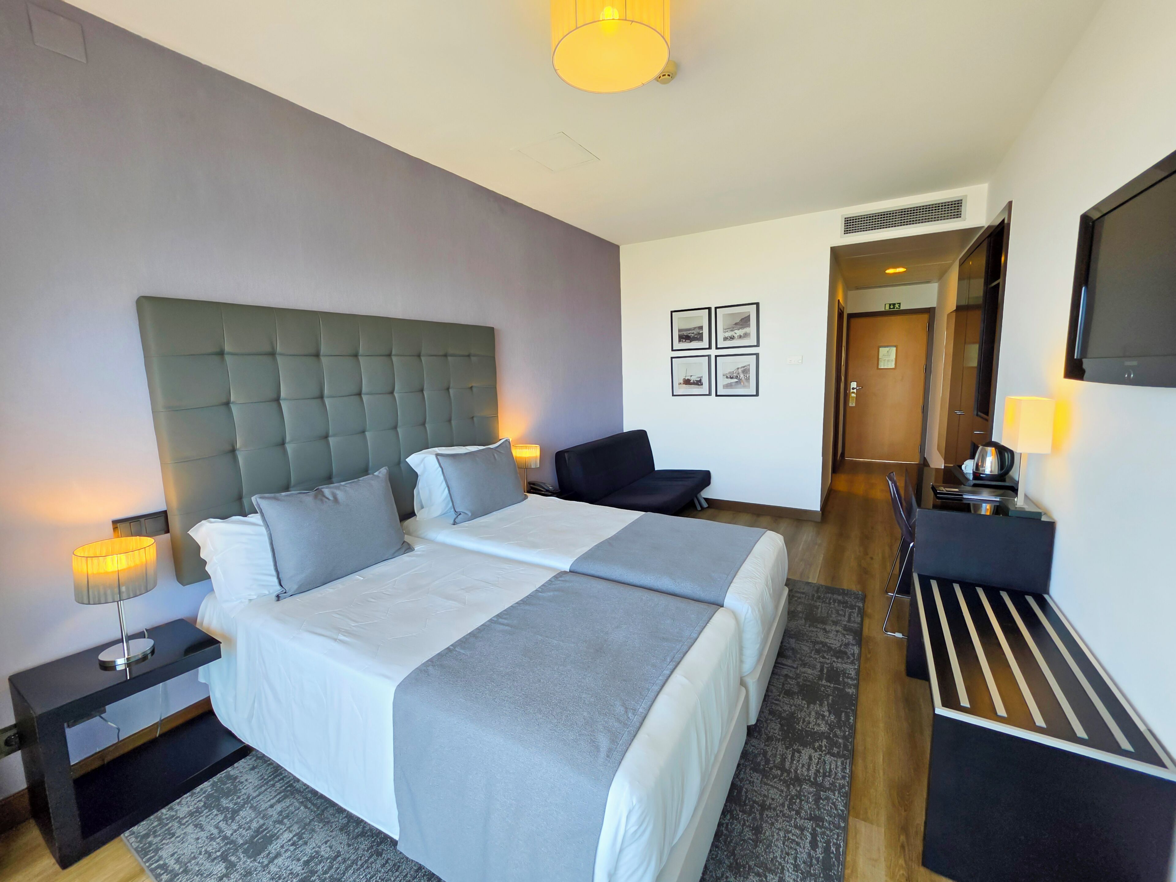 Una habitación de hotel con una cama grande, mesitas de noche, un escritorio, una silla, un televisor y cuadros en la pared.