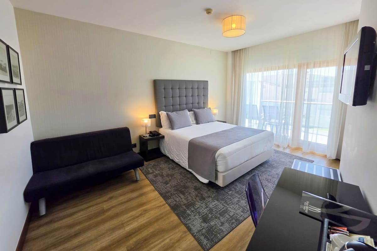 A hotel room with a bed, a sofa, a desk, a lamp, and a TV.