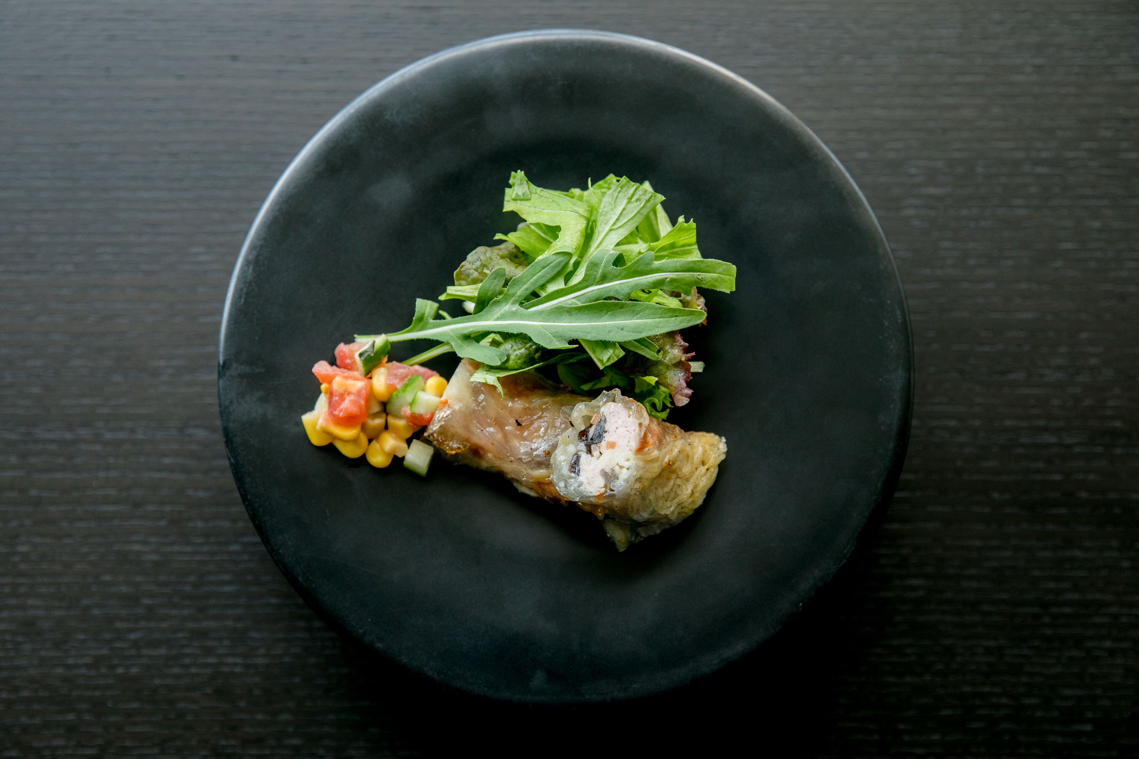 A black plate with a piece of meat, greens, and a small salad on a dark surface.
