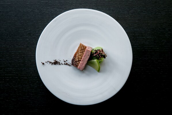 A white plate with a piece of meat and a green vegetable on a dark surface.