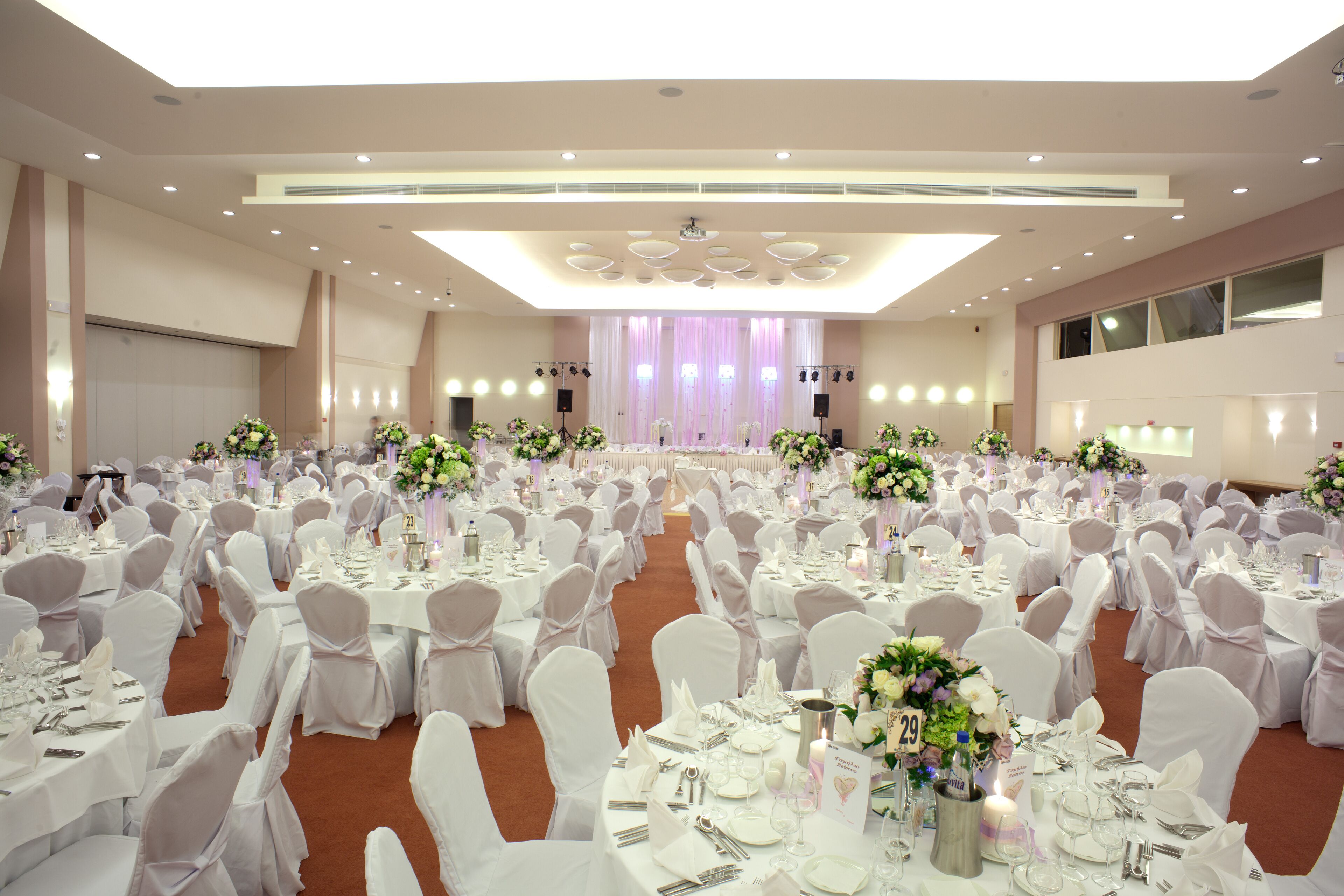 Un salón de banquetes con mesas redondas, sillas blancas y centros de mesa florales.