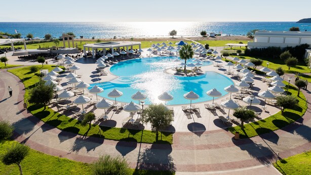 A large circular pool with surrounding lounge areas and umbrellas, set against a backdrop of a beach and ocean. - Apollo Blue (Rhodes)
