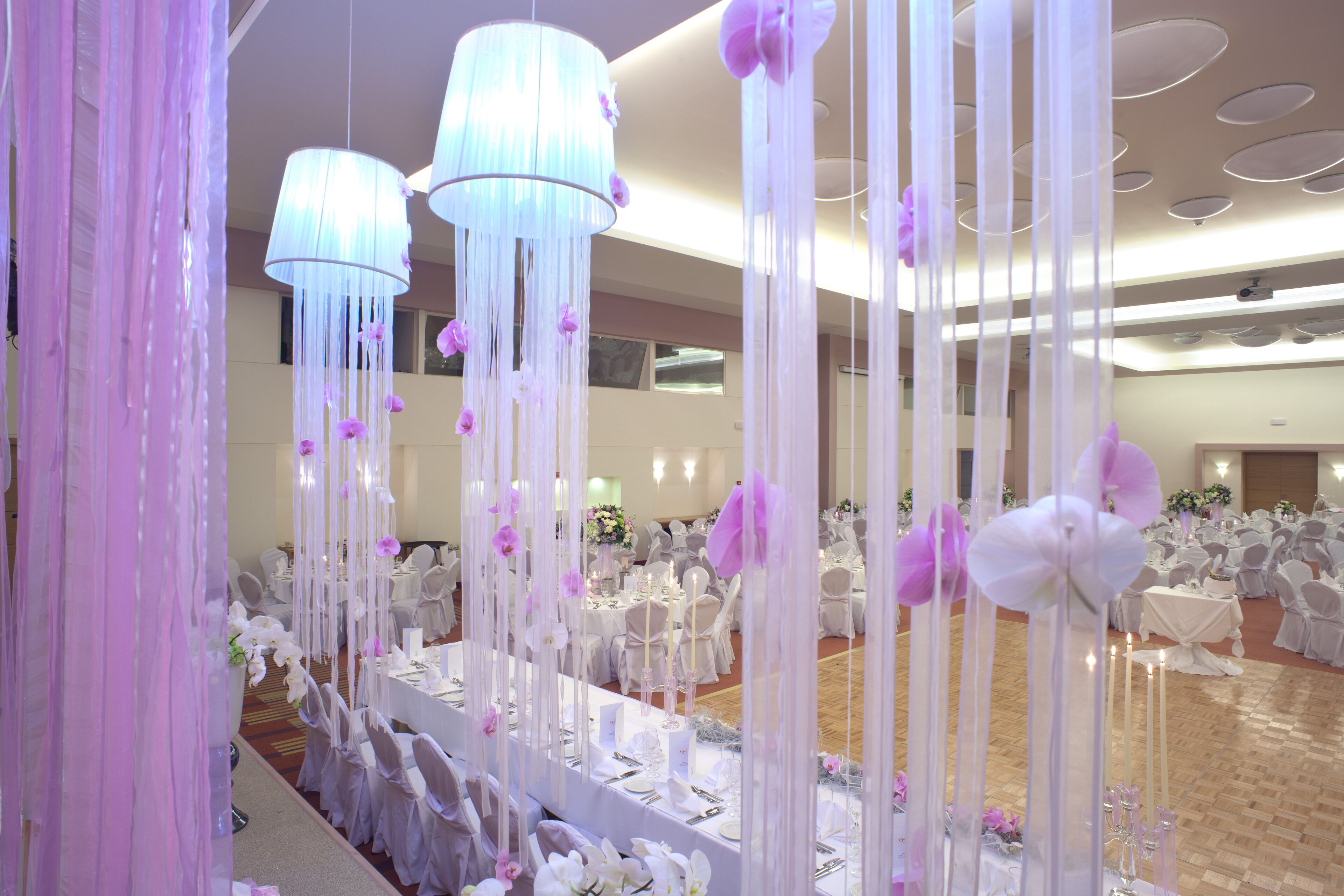 Un salón de banquetes con mesas dispuestas para un evento formal, destacando manteles blancos, centros de mesa florales y decoraciones florales colgantes.