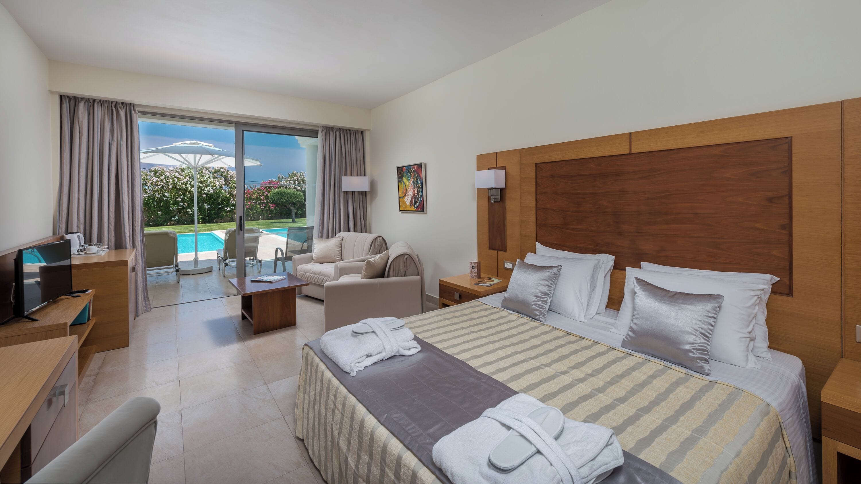 Une chambre d’hôtel avec un grand lit, un bureau, une télévision, un canapé, une table basse et une vue sur une piscine.