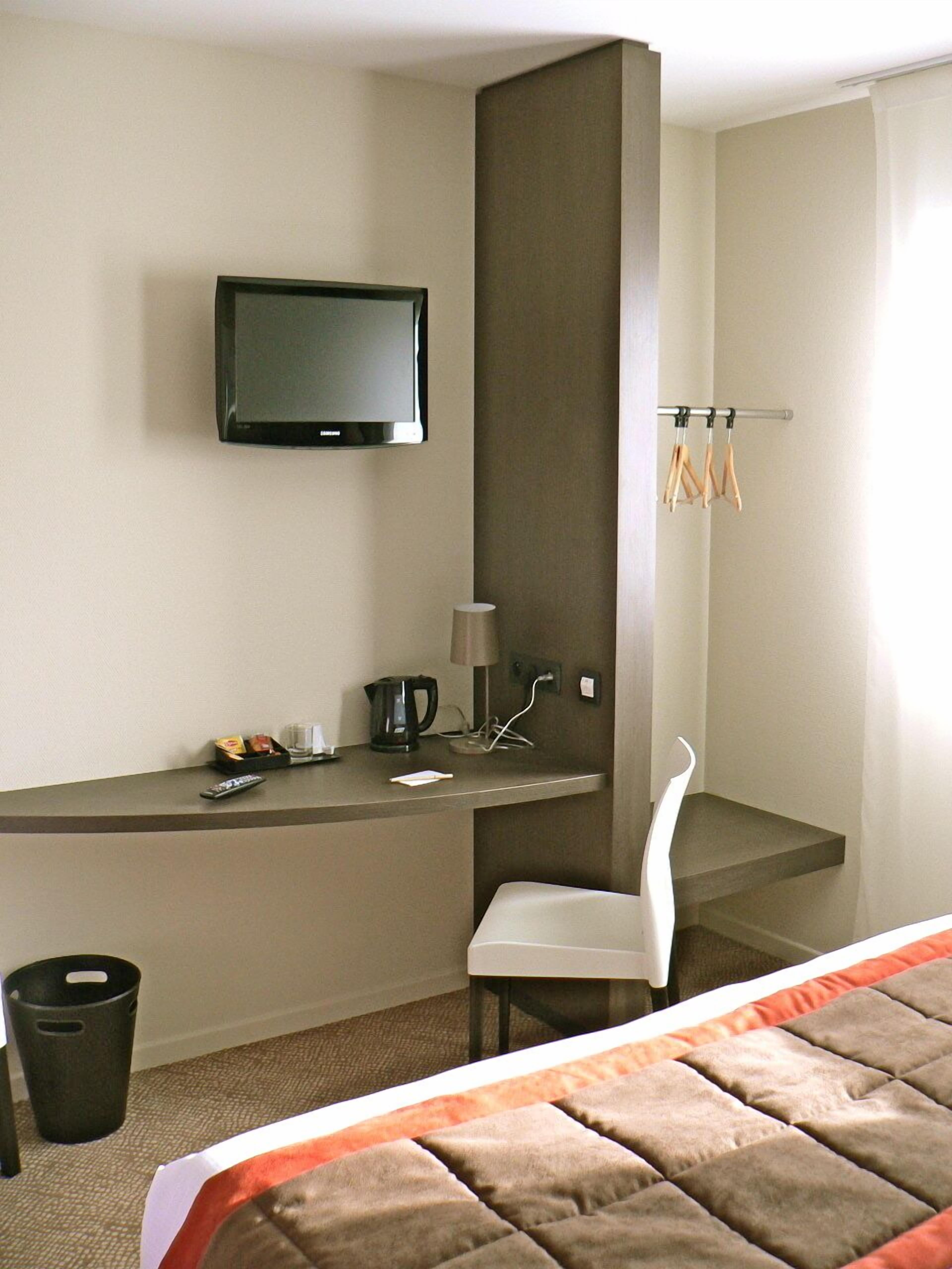 A hotel room with a bed, a desk with a TV, a chair, a coffee maker, and a trash bin.
