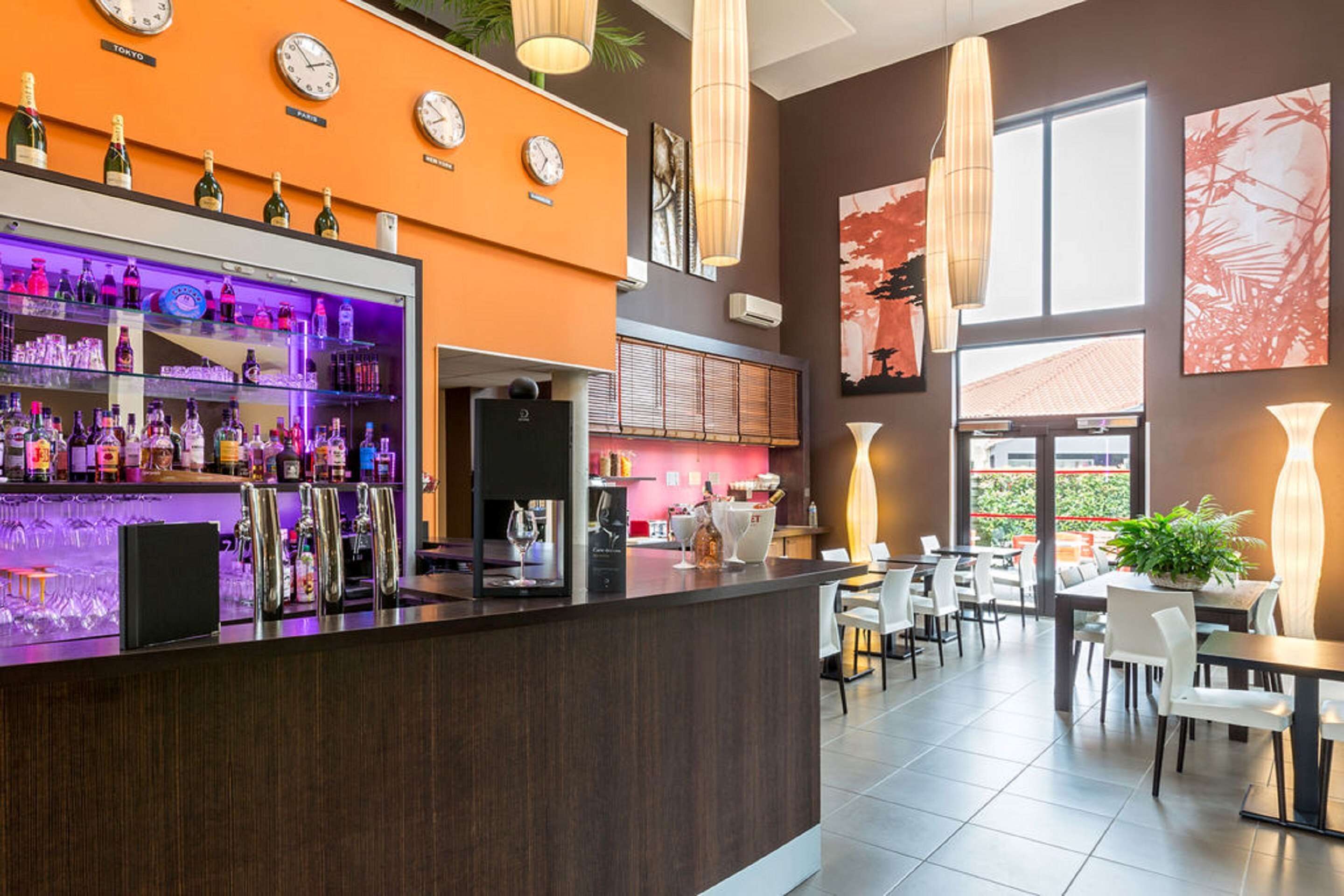 Una zona de bar con varios relojes, una barra llena de botellas y asientos en una sala amplia con ventanas grandes.