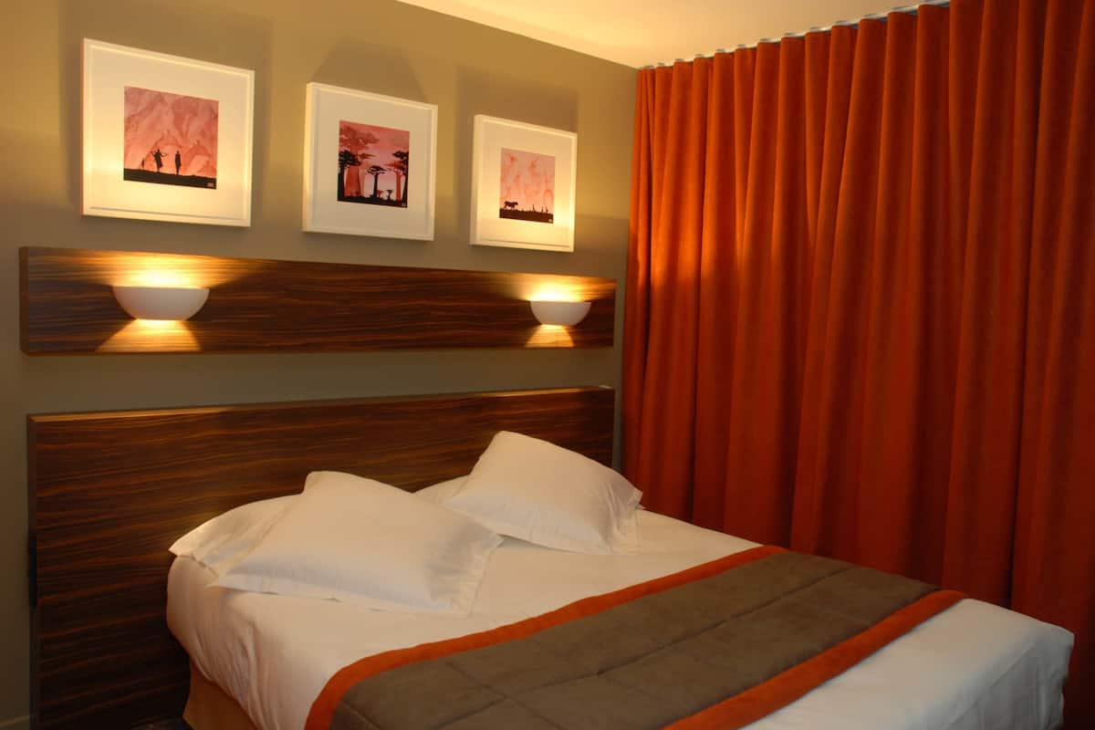 Un lit bien fait, avec du linge de lit blanc et un plaid marron assorti, une tête de lit en bois et trois tableaux encadrés accrochés au mur.