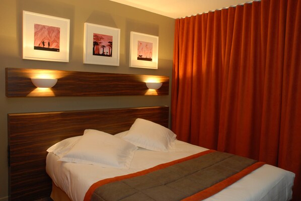 A neatly made bed with white linens and a brown accent throw, a wooden headboard, and three framed pictures on the wall.