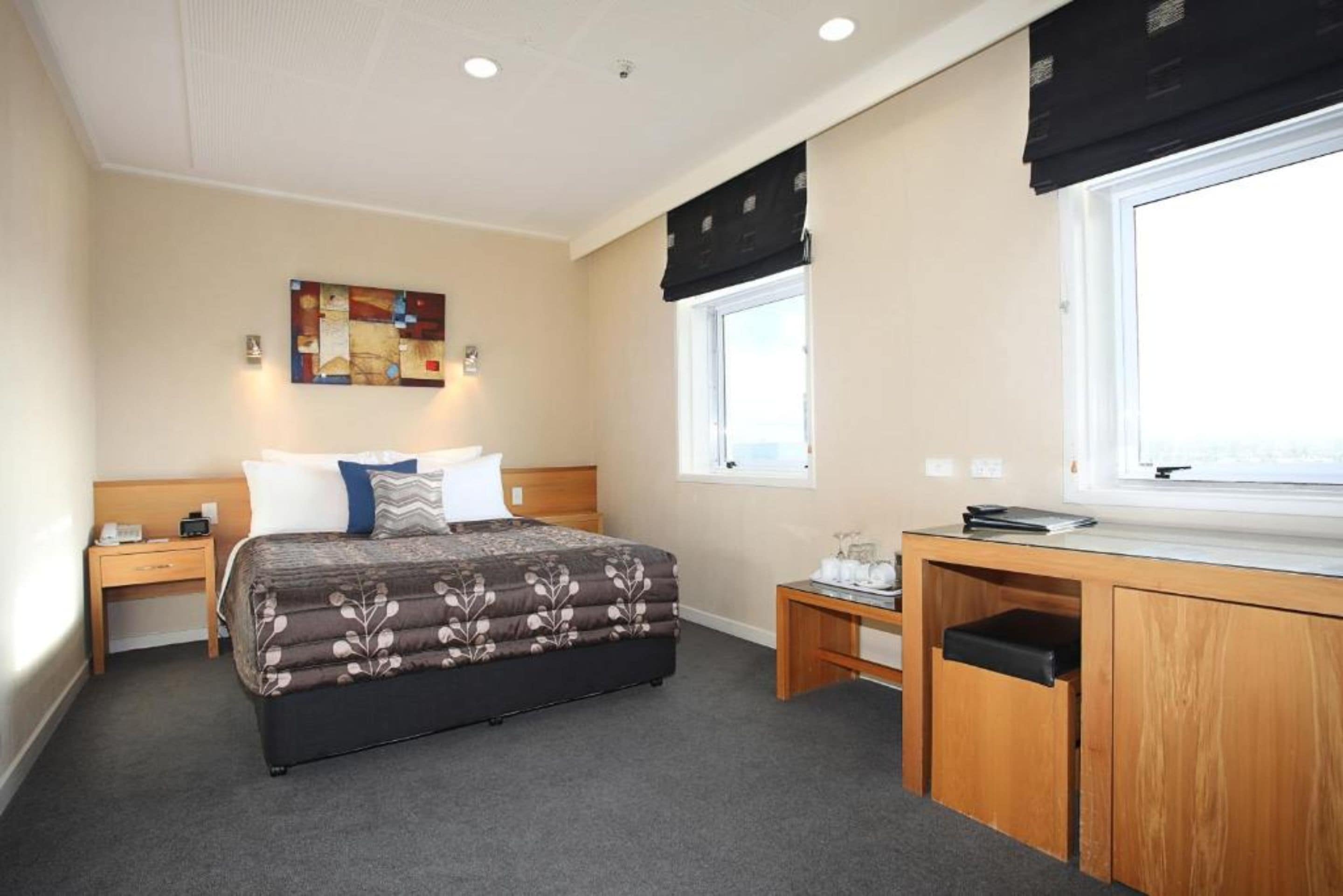 A hotel room with a bed, a desk, a chair, a nightstand, a lamp, a window with a view, and a painting on the wall.