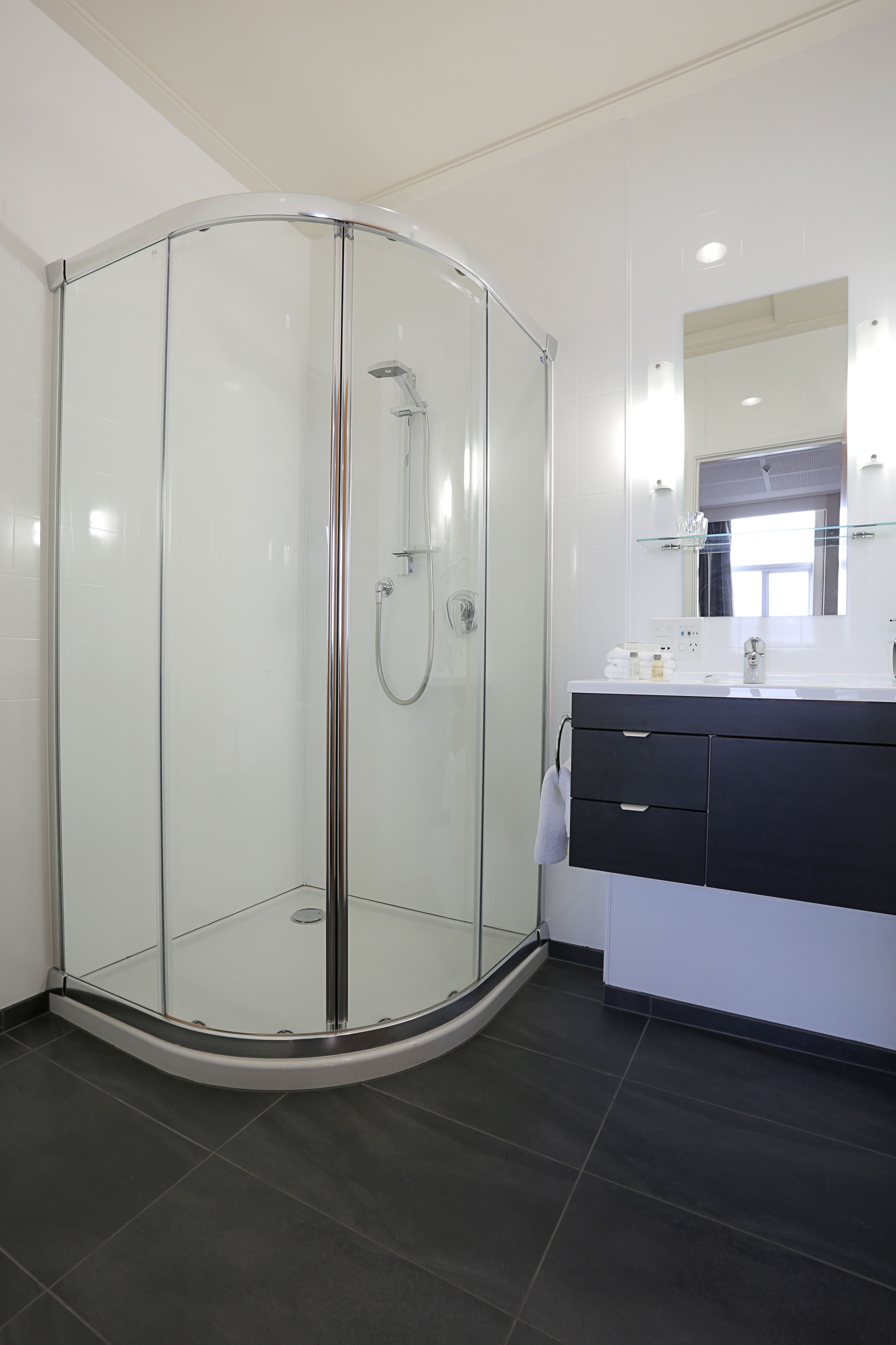 A modern bathroom with a glass-enclosed shower, a dark-tiled floor, and a white vanity with a mirror.