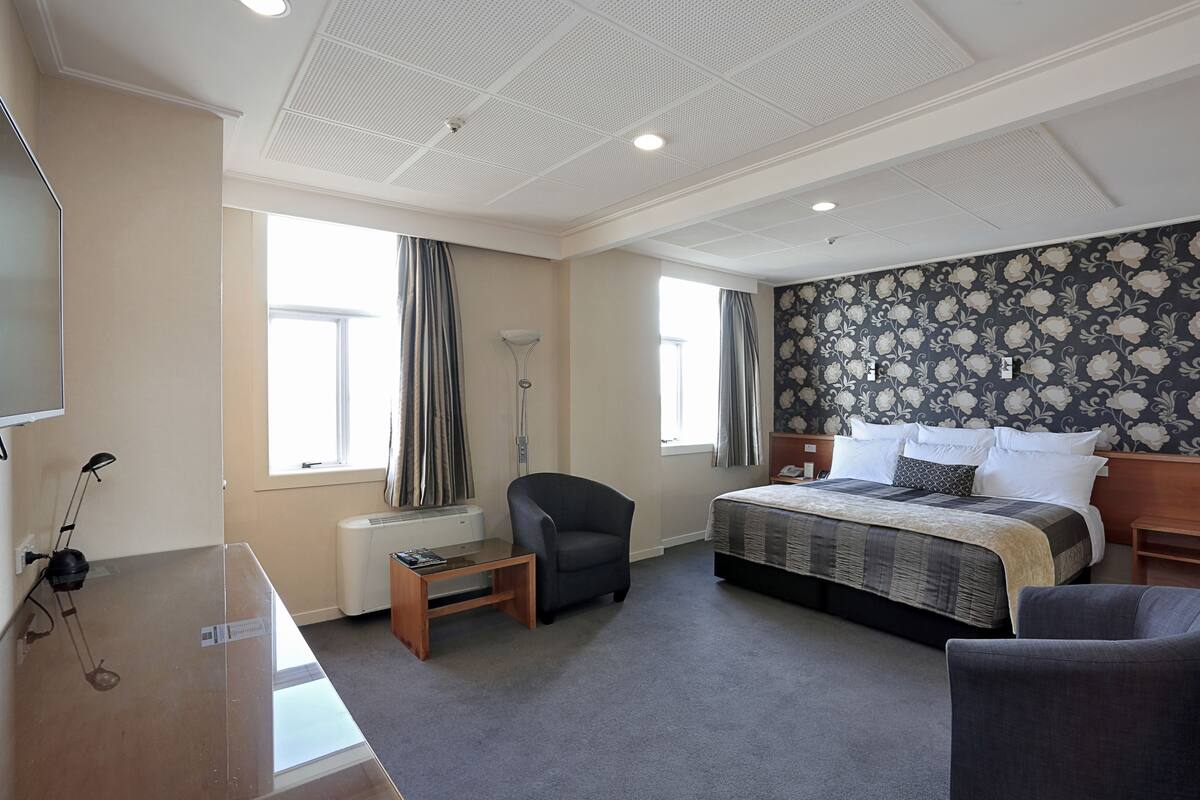 Quarto de hotel com cama, escrivaninha, cadeira, abajur e uma grande janela.