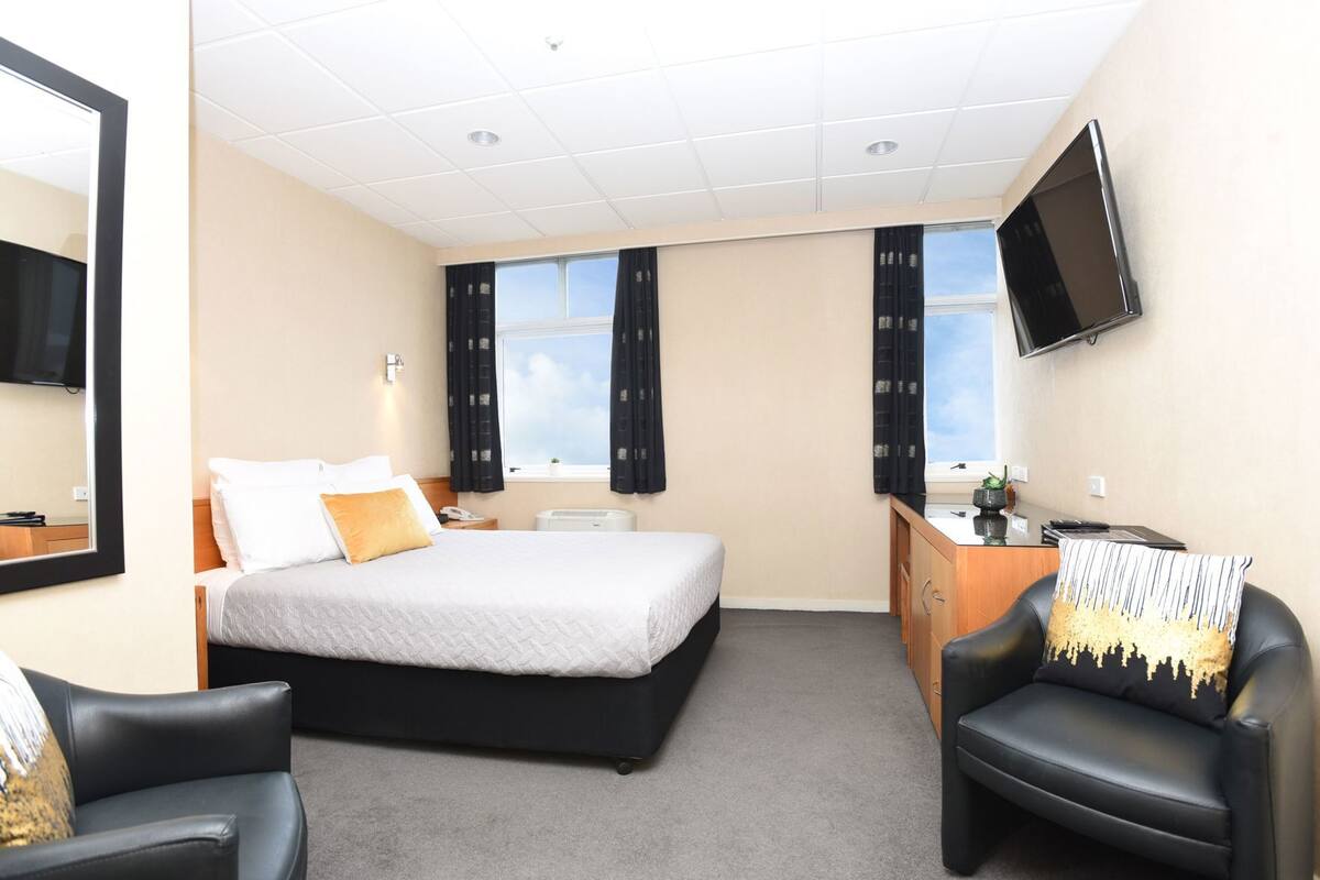 Quarto de hotel com cama, escrivaninha, cadeira, televisão e janela com cortinas.