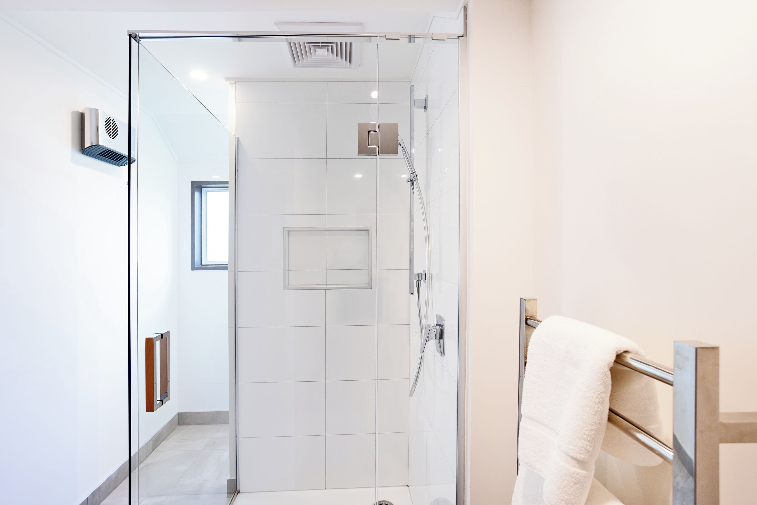 A modern bathroom with a glass shower enclosure, a towel rack, and a visible ventilation system.