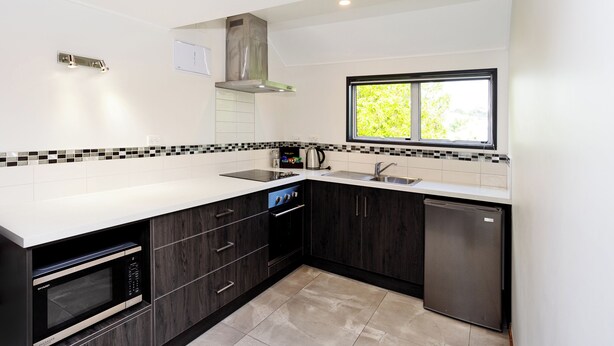 A modern kitchen with dark wood cabinets, a stainless steel dishwasher, a built-in oven, a microwave, and a window providing natural light. - Balmoral Lodge Motel (Invercargill)
