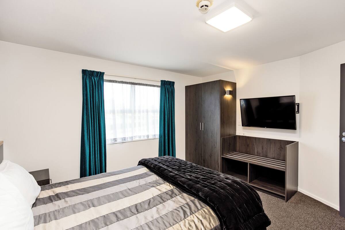 A hotel room with a bed, a TV mounted on the wall, a wardrobe, and a window with curtains.