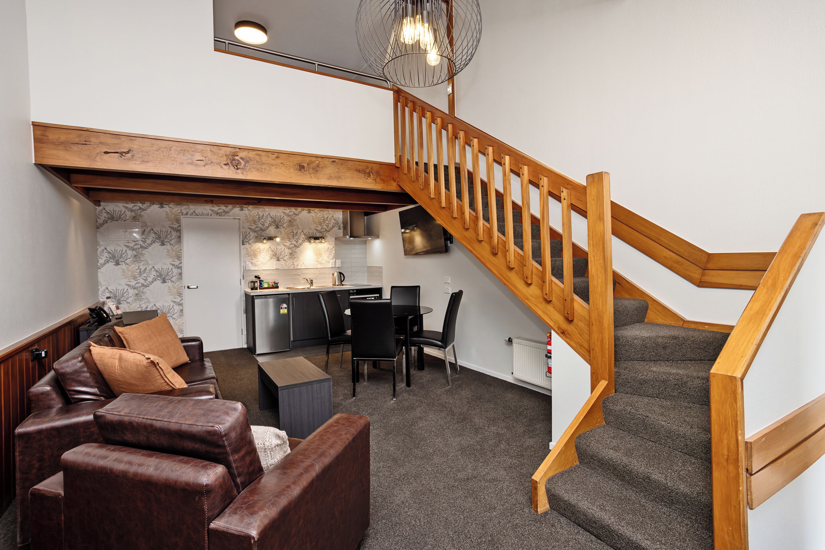 Una sala de estar moderna con un sofá de cuero marrón, una mesa de comedor con sillas, una zona de cocina y una escalera de madera.