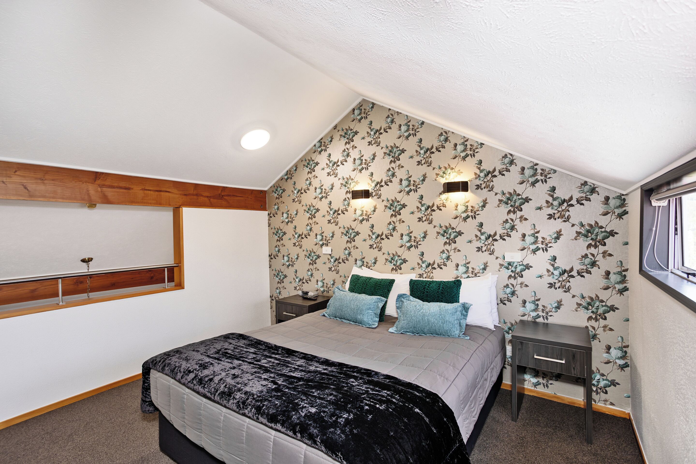 A bedroom with a sloped ceiling, floral wallpaper, a bed with a black and white duvet, two bedside tables, and a window with a view.