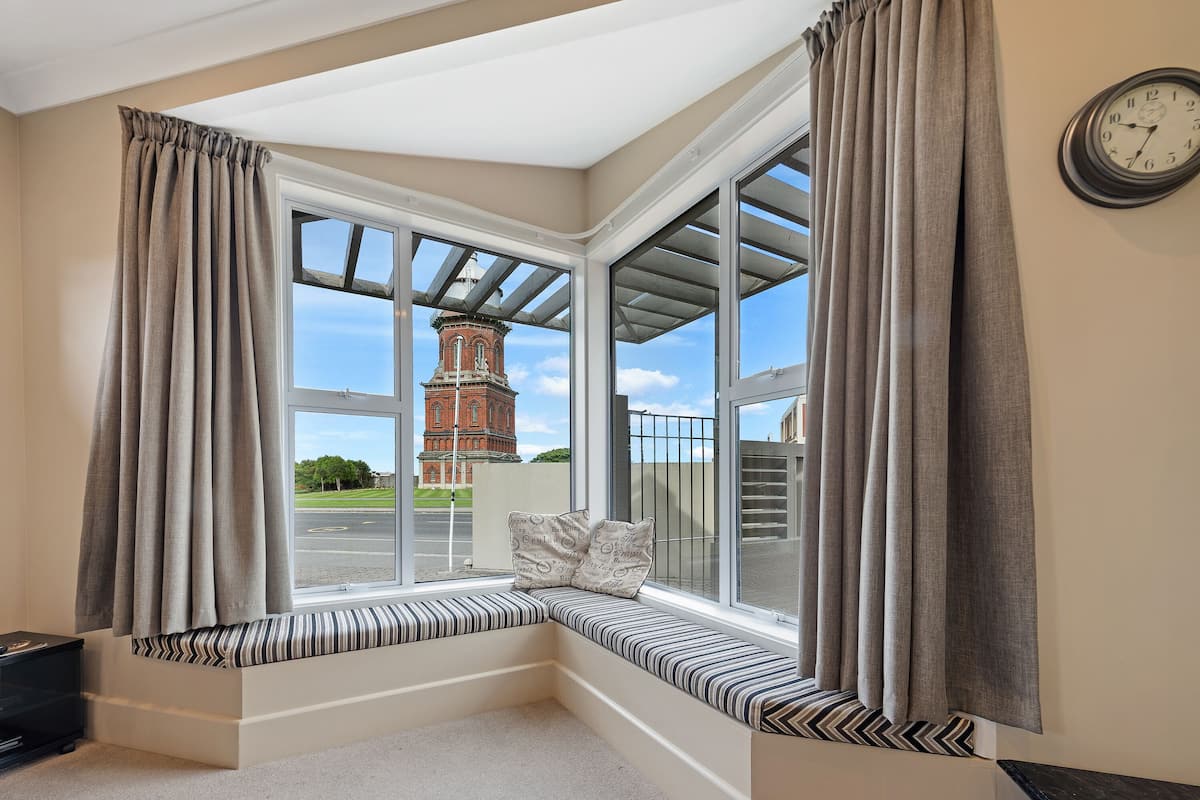 A corner window seat with a bench, curtains, and a clock on the wall.