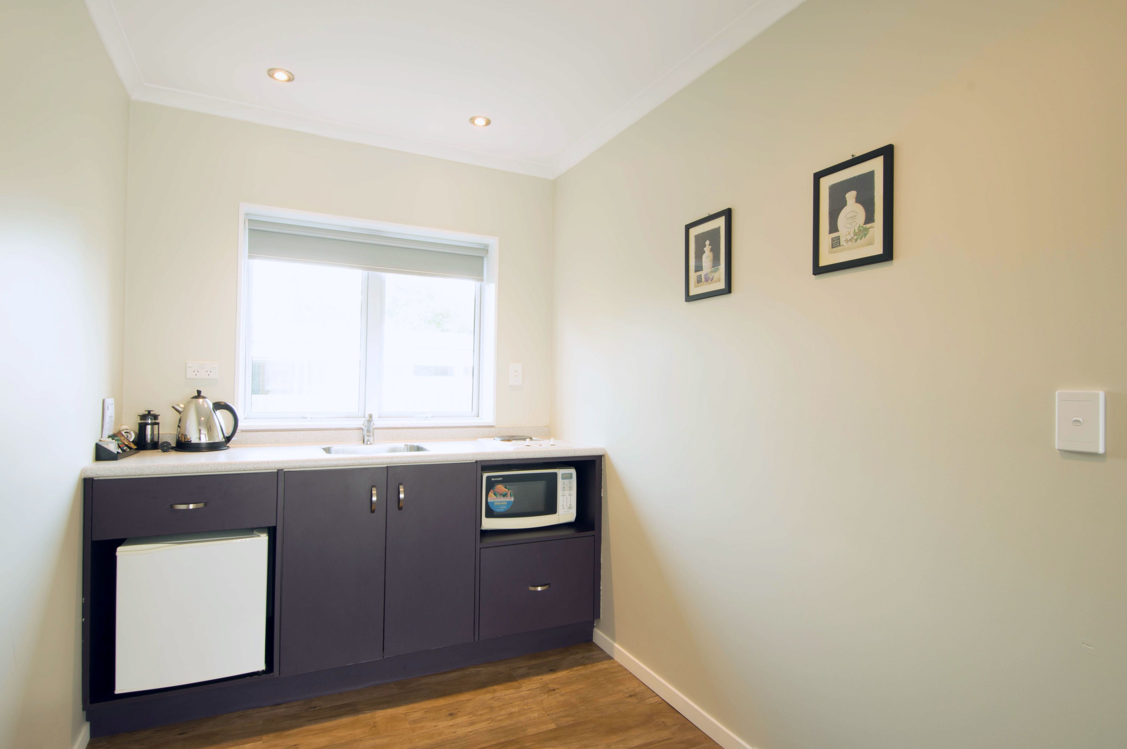 A compact kitchen with a refrigerator, microwave, and kettle on the counter, a window above the sink, and two framed pictures on the wall.