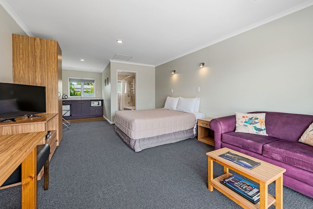 A modern hotel room with a bed, a TV, a purple sofa, and a small wooden table.