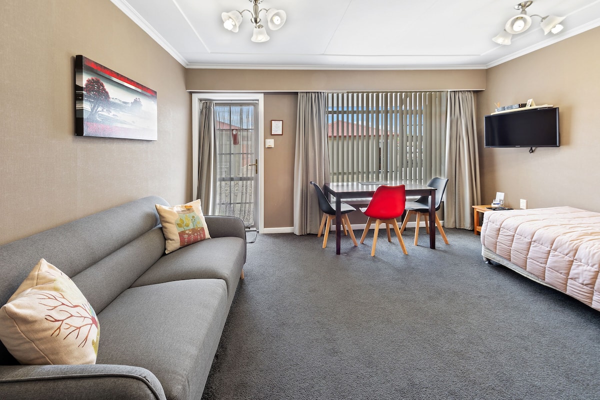 A hotel room with a grey sofa, a red chair, a bed, a desk with a chair, and a TV mounted on the wall.