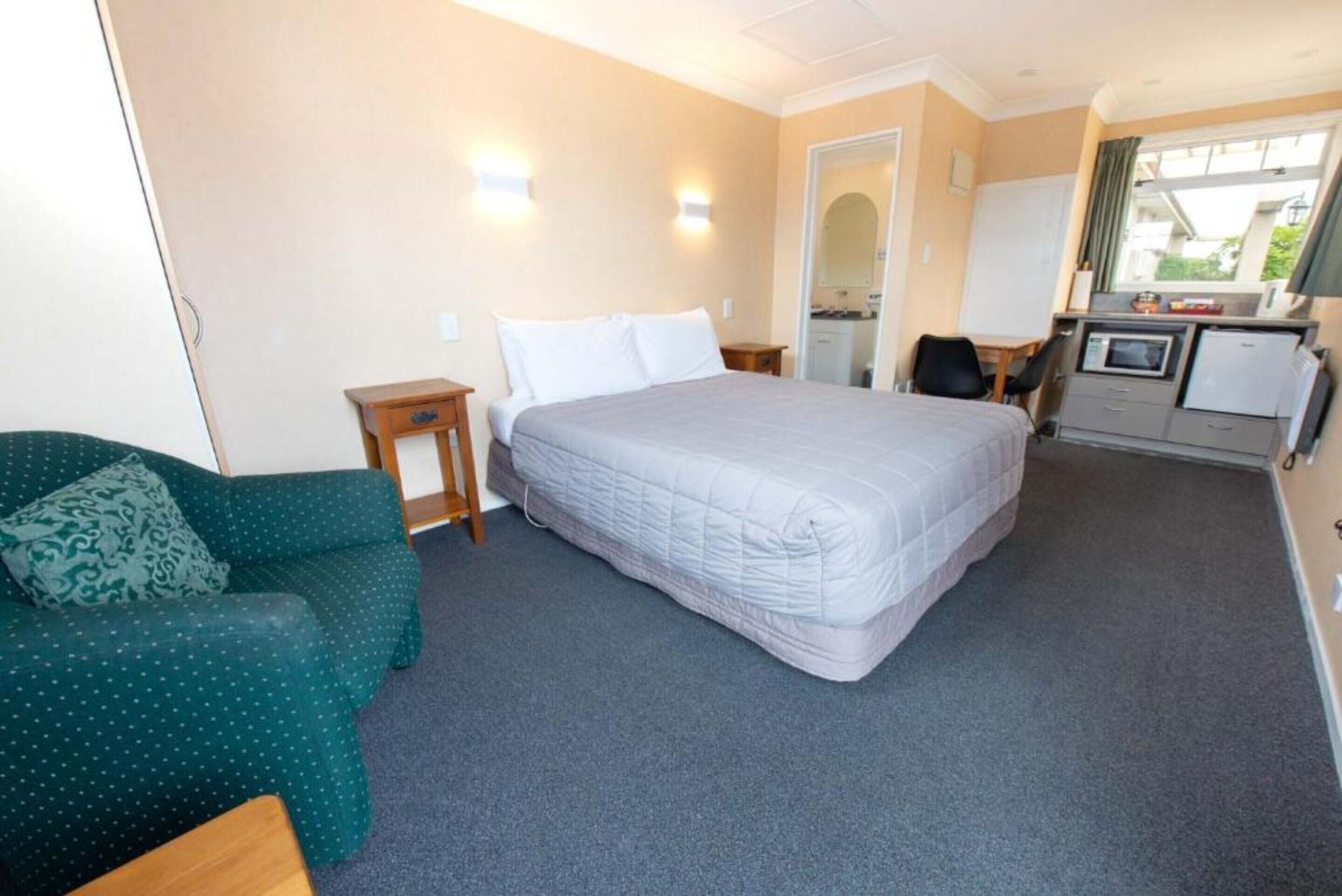A hotel room with a bed, a green sofa, a small table, a desk with a computer, and a kitchenette.
