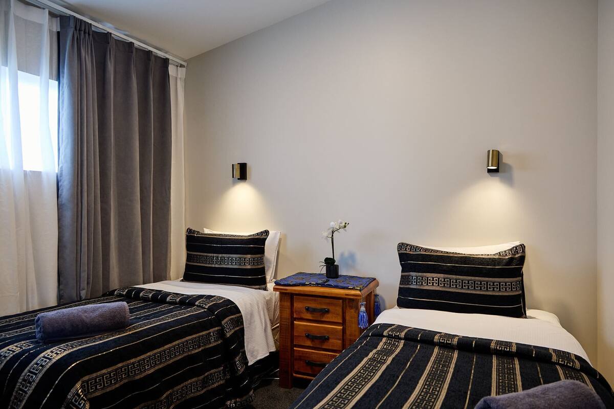 Quarto de hotel com duas camas, cada uma com roupa de cama estampada, uma pequena mesa de cabeceira de madeira com um vaso de flores e luminárias fixadas na parede.