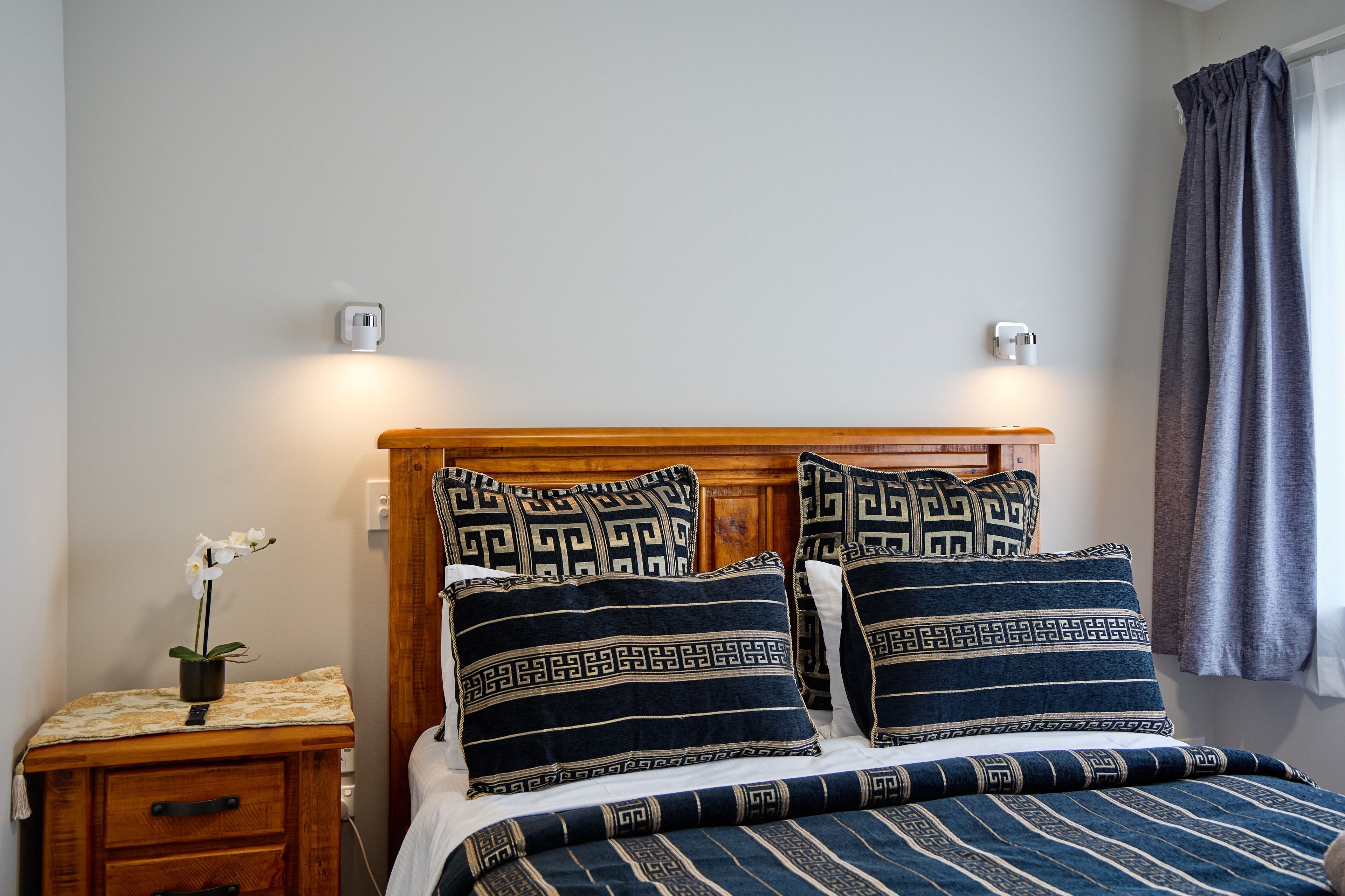 A neatly made bed with patterned pillows and a wooden headboard, a bedside table with a plant, and a power outlet on the wall.