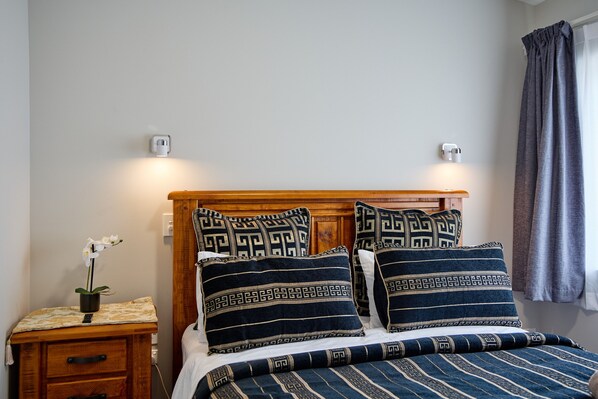 A neatly made bed with patterned pillows and a wooden headboard, a bedside table with a plant, and a power outlet on the wall. - Willowbank Motel (Kaikoura)