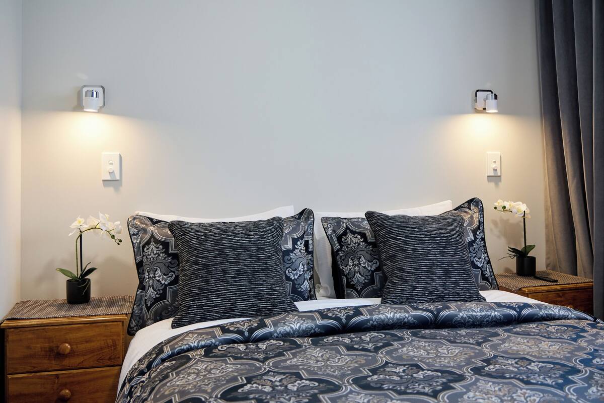 Uma cama bem arrumada com roupa de cama estampada, duas mesas de cabeceira com abajures e orquídeas em vasos de cada lado.