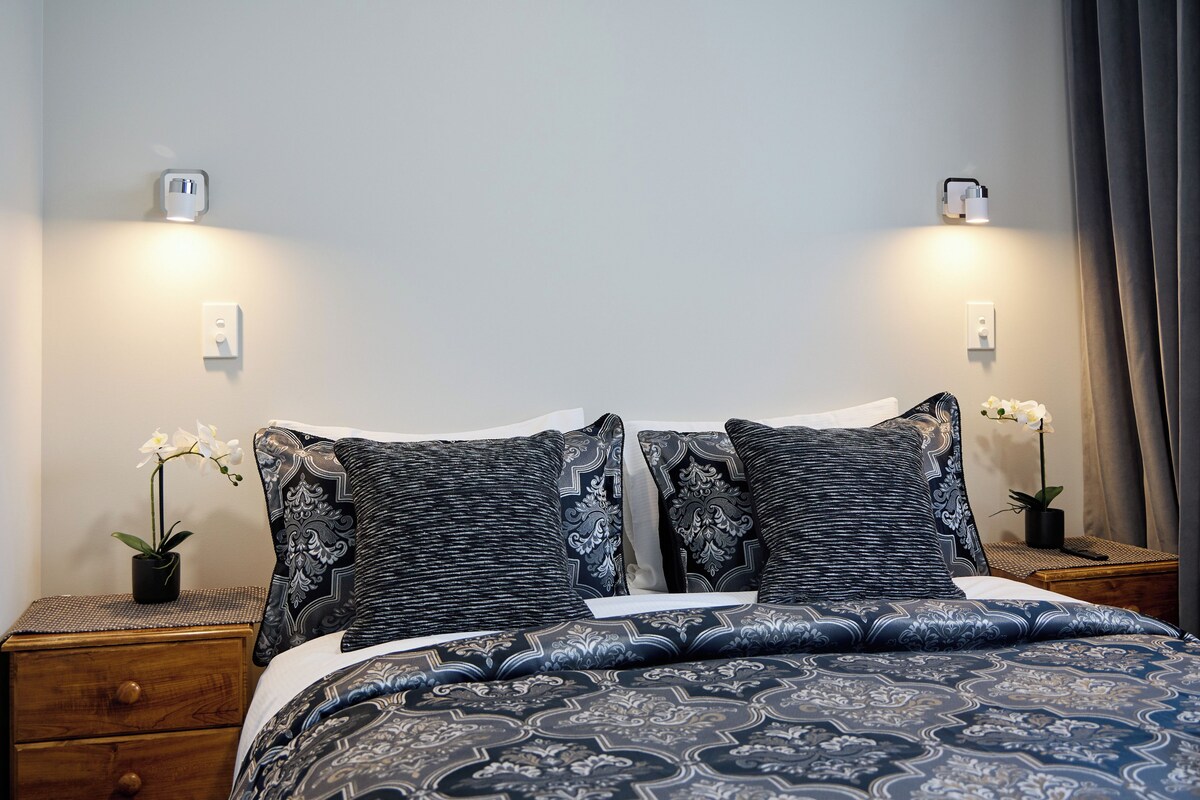 Una cama bien hecha con ropa de cama estampada, dos mesitas de noche con lámparas y orquídeas en macetas a cada lado.