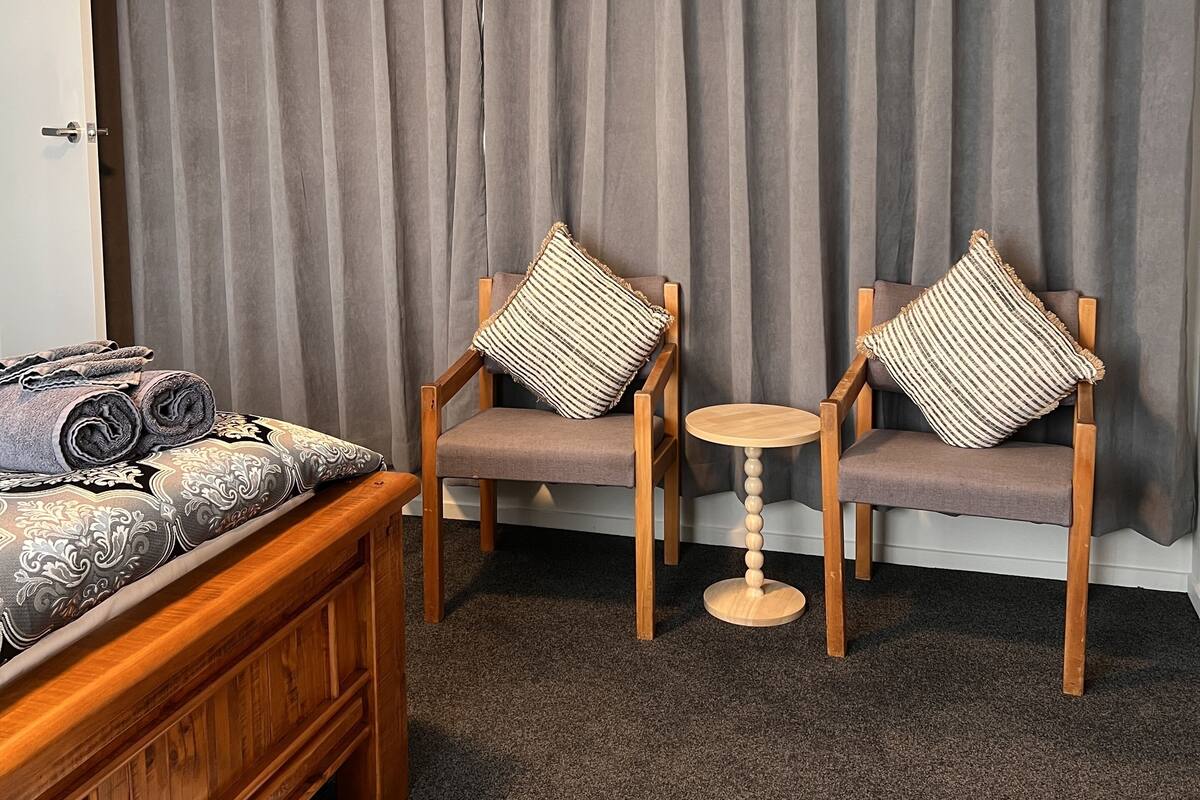 Quarto com cama de madeira, duas cadeiras de madeira com almofadas, uma mesa redonda pequena e um painel de cortina cinza.
