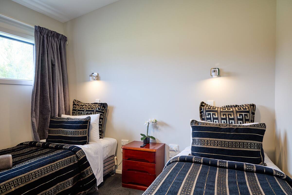 Quarto com duas camas, cada uma com roupa de cama estampada, uma mesa de cabeceira com abajur e uma janela com cortinas.