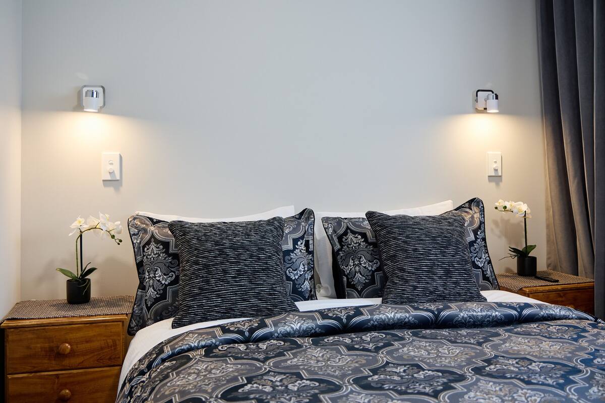 Uma cama bem arrumada com roupa de cama estampada, duas mesas de cabeceira com abajures e orquídeas em vasos de cada lado.