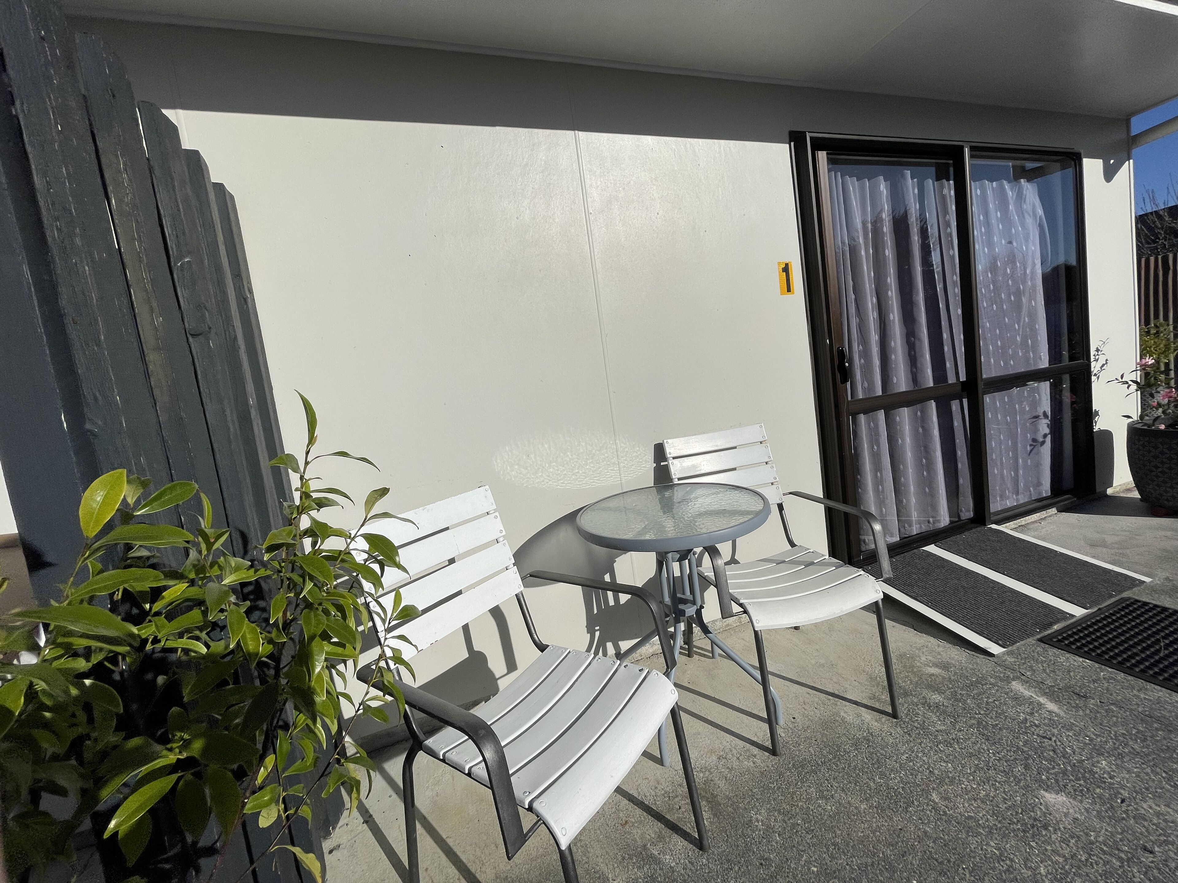 Un patio con un banco blanco, una mesa redonda, una planta en maceta y un ventanal corredero con cortinas.