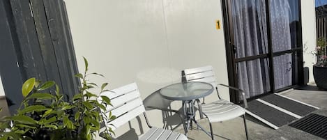 Un patio con un banco blanco, una mesa redonda, una planta en maceta y un ventanal corredero con cortinas.