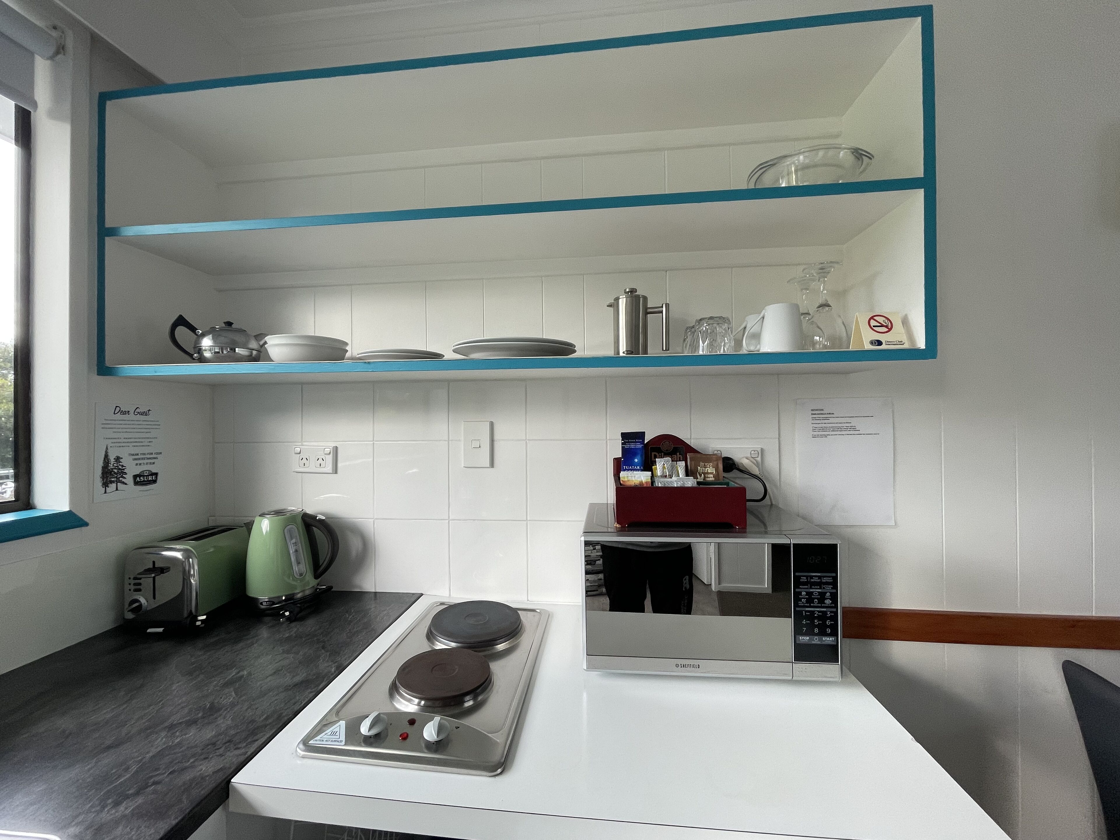 A kitchen with a stove, microwave, and toaster. There is a shelf above the stove with dishes and a kettle.