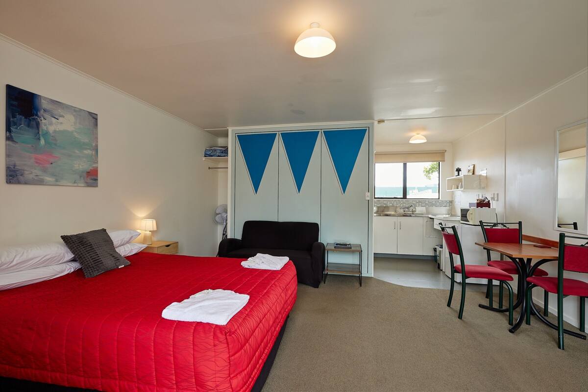 Quarto de hotel com cama vermelha, uma escrivaninha e uma cadeira. É visível uma kitchenette ao fundo.