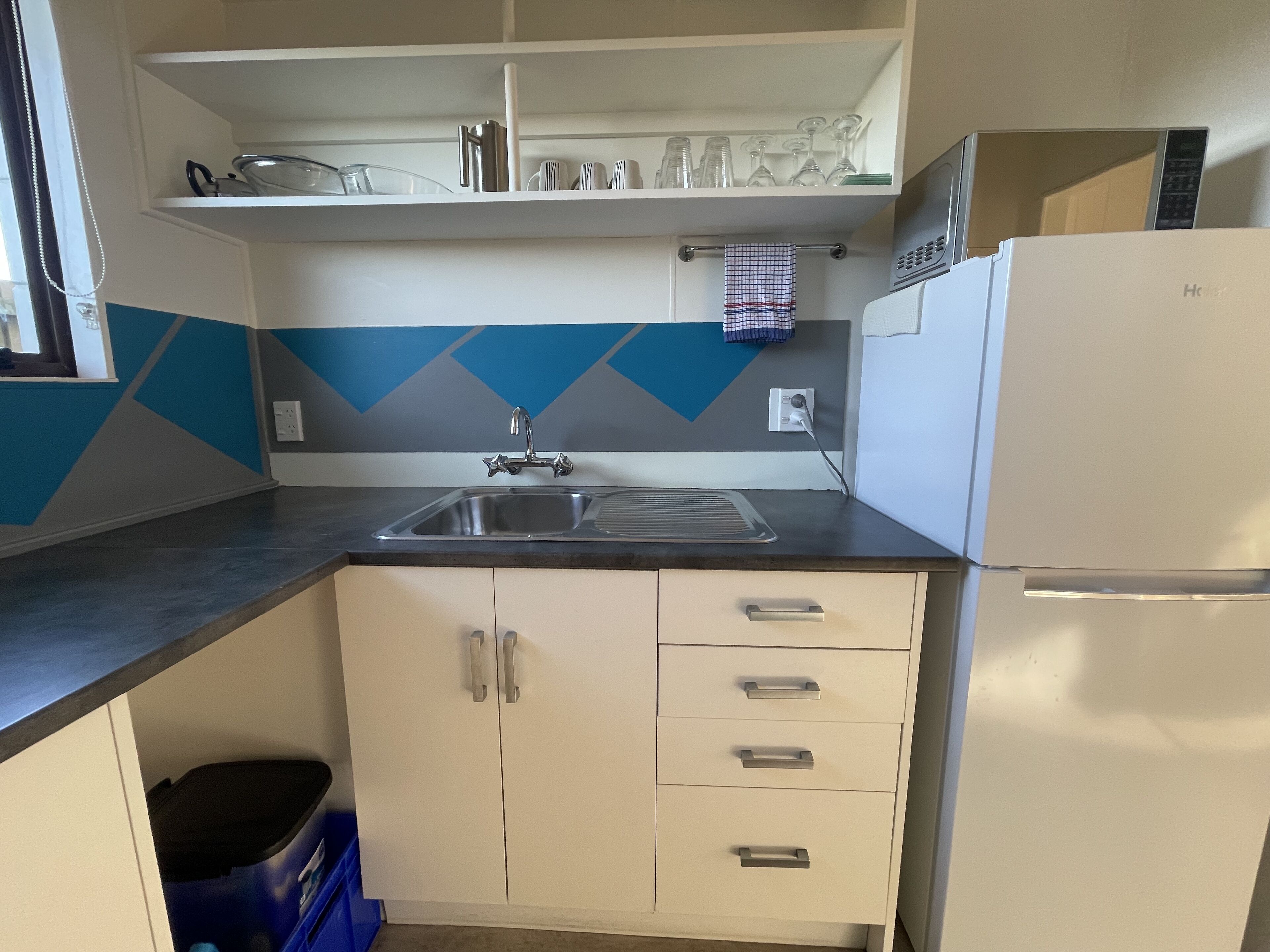 Cocina compacta con lavamanos, refrigerador y encimera. Hay armarios con cajones y un cubo de basura azul.