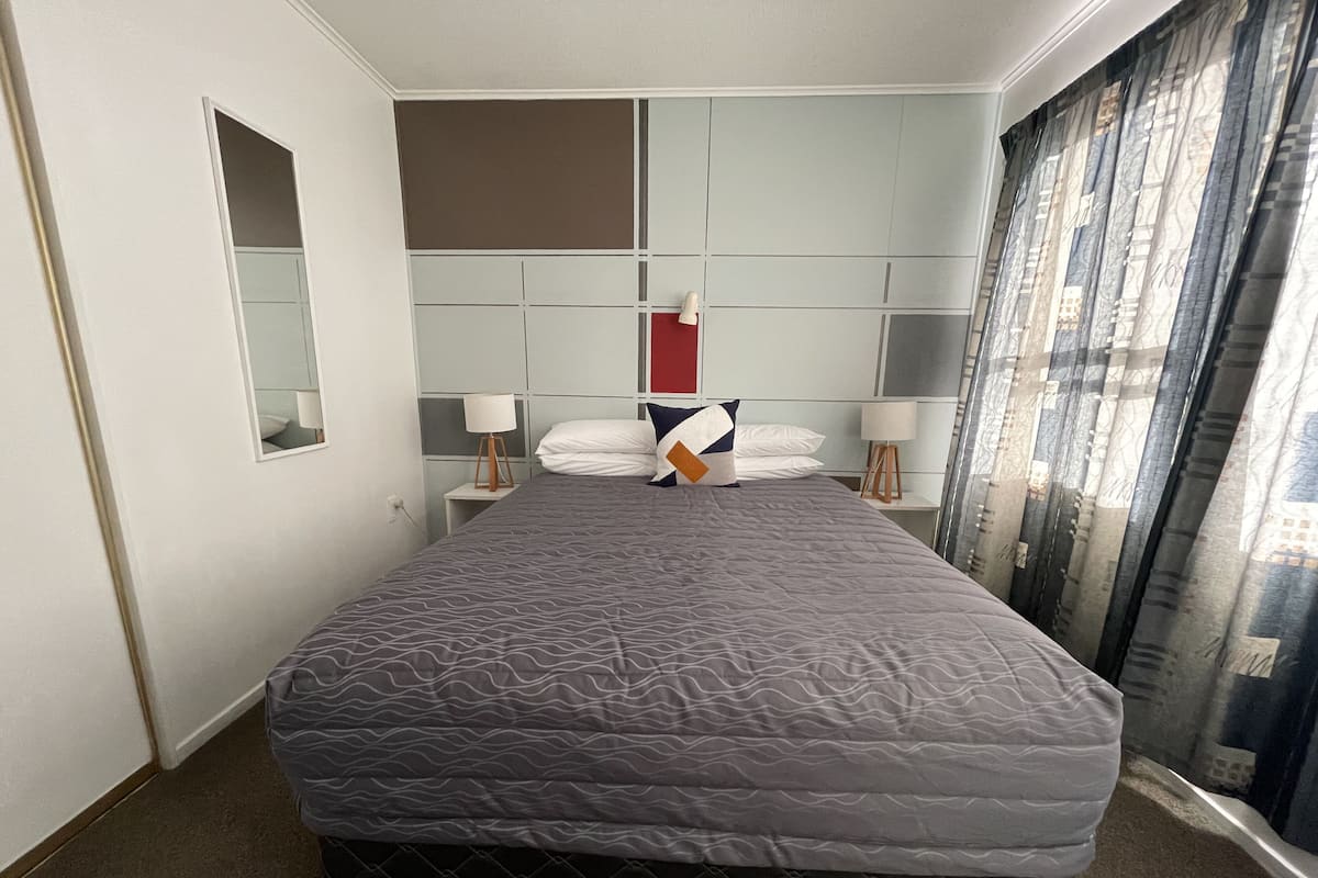 Quarto de hotel com uma cama grande, mesa de cabeceira com abajur, espelho e vista da cidade através das cortinas.
