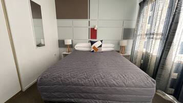 Habitación de hotel con una cama grande, una mesita de noche con lámpara, un espejo y vistas a la ciudad a través de cortinas.