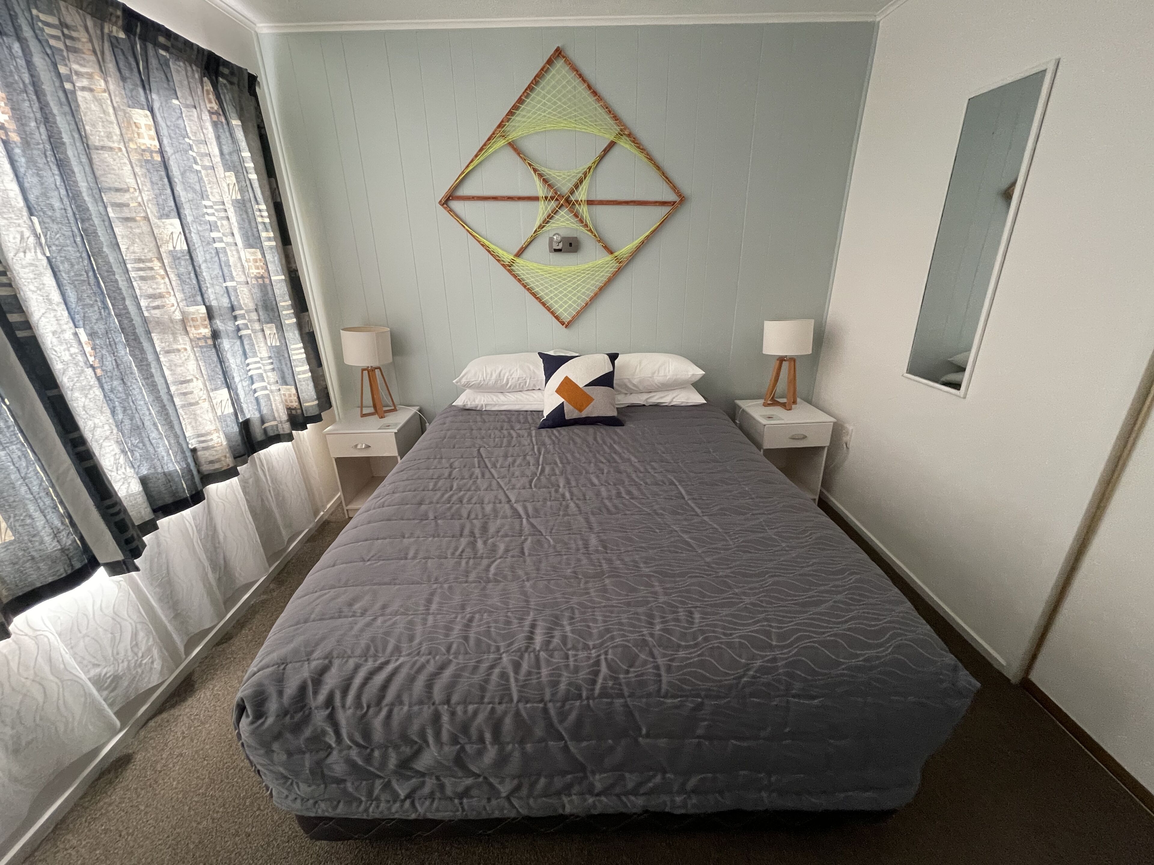A hotel room with a large bed, two bedside tables with lamps, a decorative wall hanging, and a window with curtains.