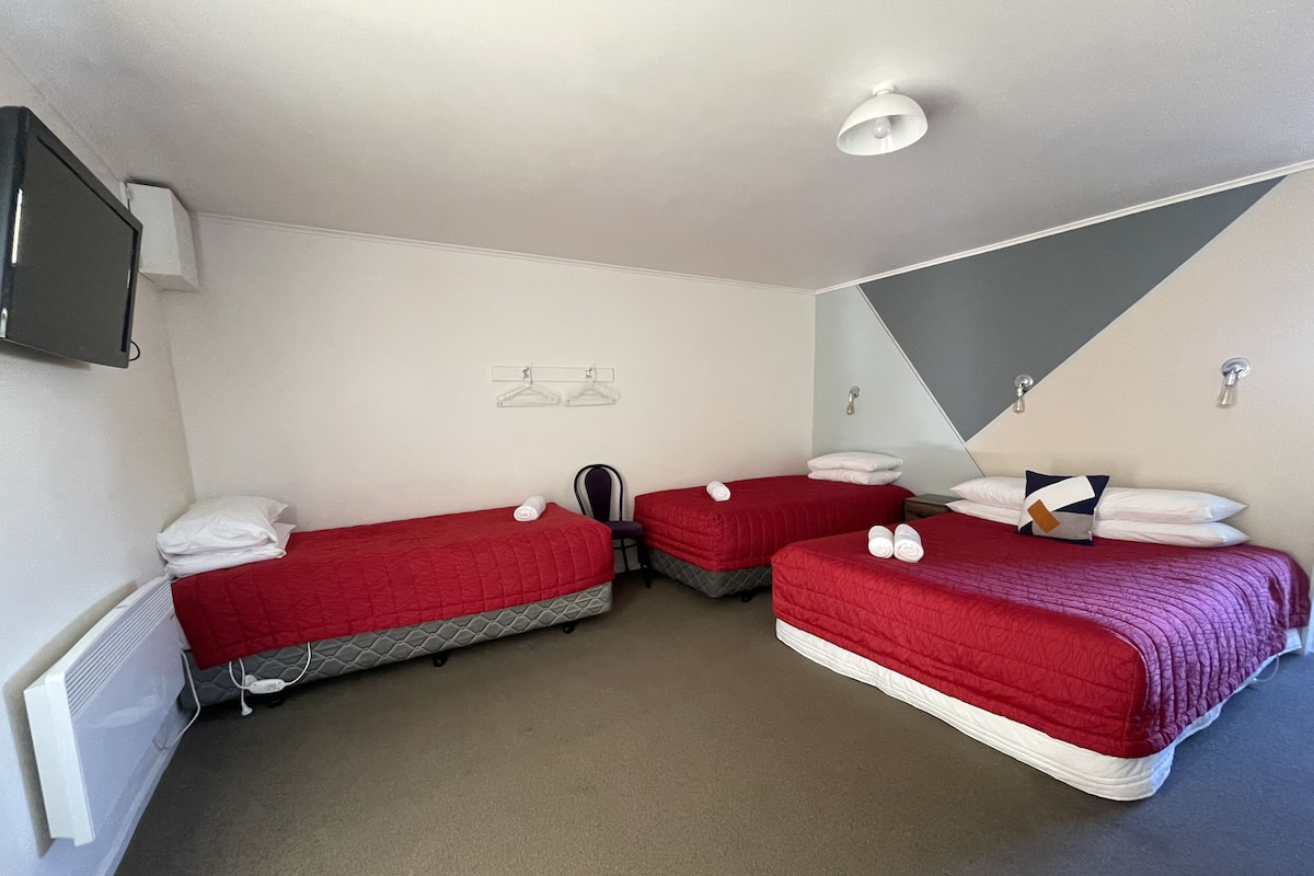 Quarto com duas camas individuais, cada uma com roupa de cama vermelha e almofadas brancas, uma cadeira pequena e um radiador.