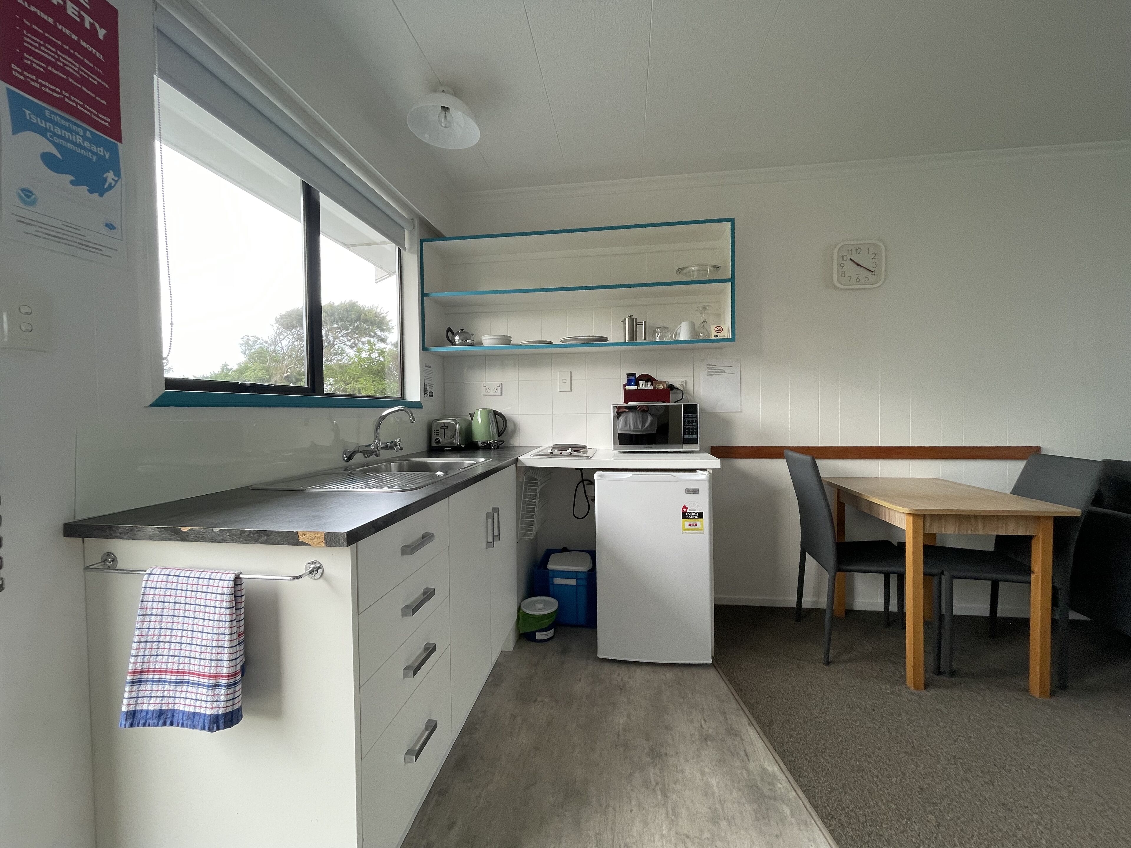 A compact kitchen with a sink, a microwave, a toaster, and a small refrigerator. There is a window providing natural light and a view of the outdoors. A table and chairs are positioned near the window.