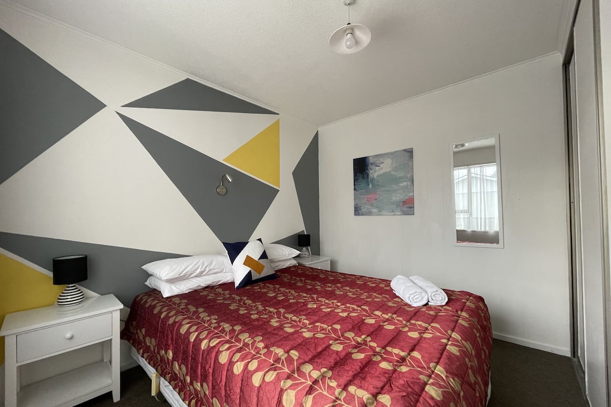 Quarto com cama, mesa de cabeceira, abajur, pintura e janela.