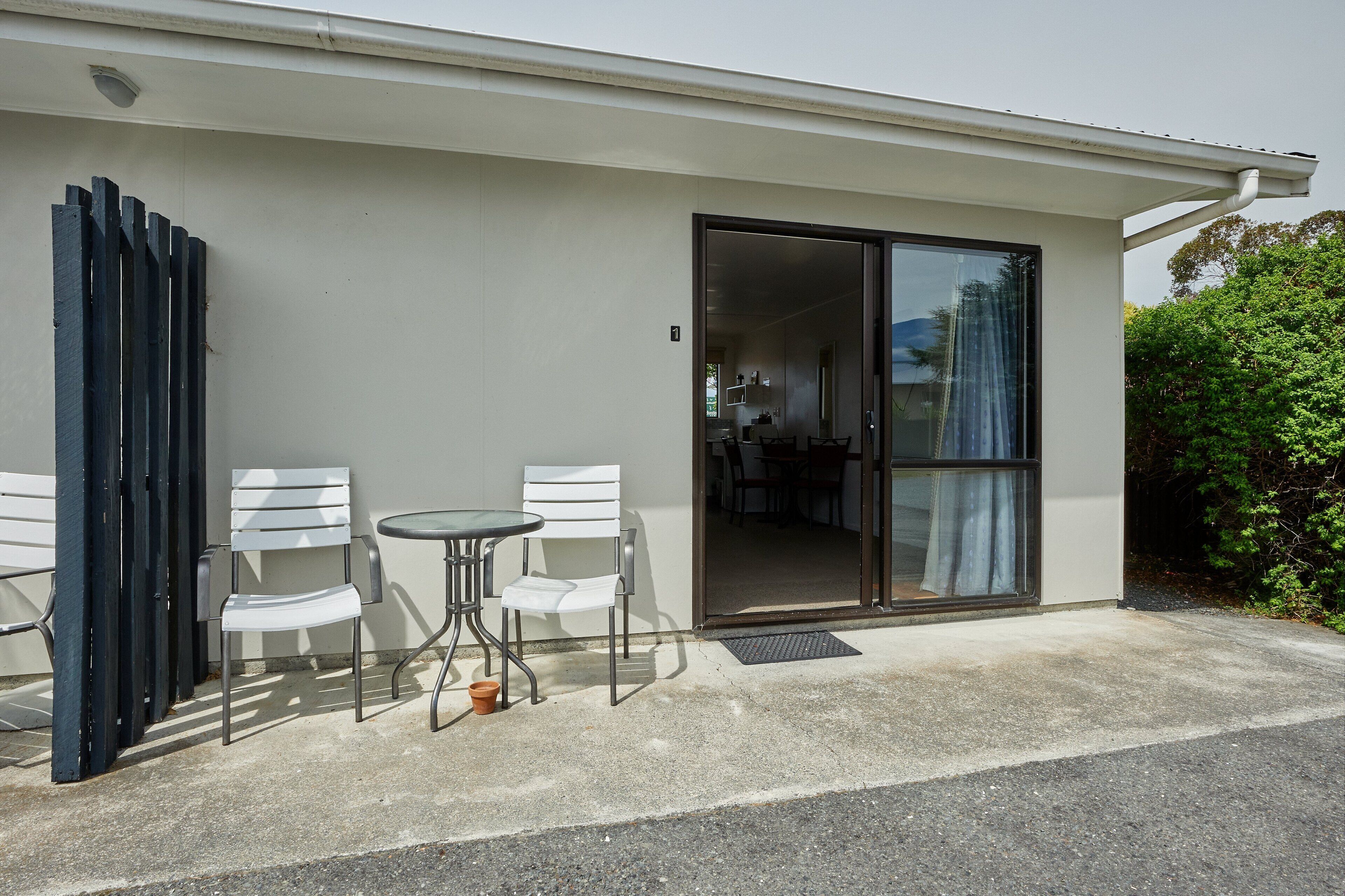 A modern building with a sliding glass door entrance, a small patio with two white chairs and a table, and a visible kitchen area inside.