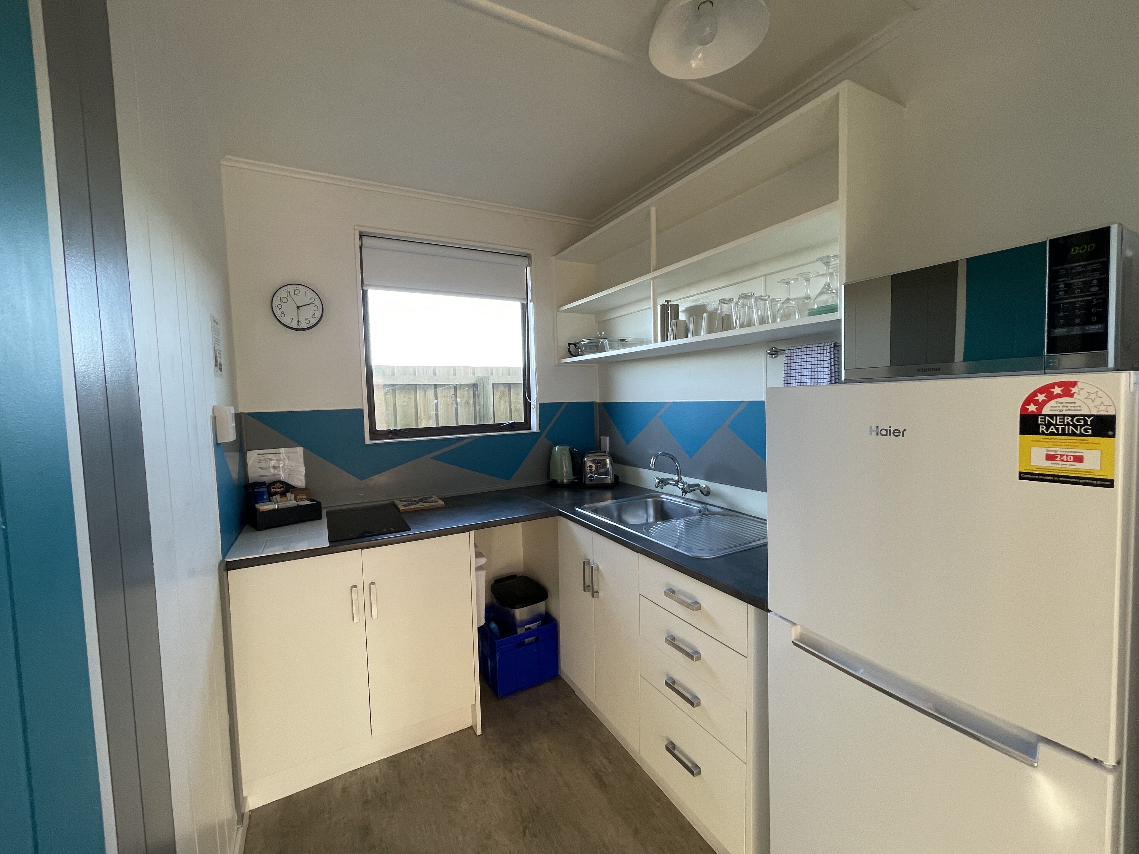 Une cuisine compacte avec des meubles blancs, un pare-feu bleu et gris, un réfrigérateur Haier et une fenêtre donnant sur une clôture en bois.
