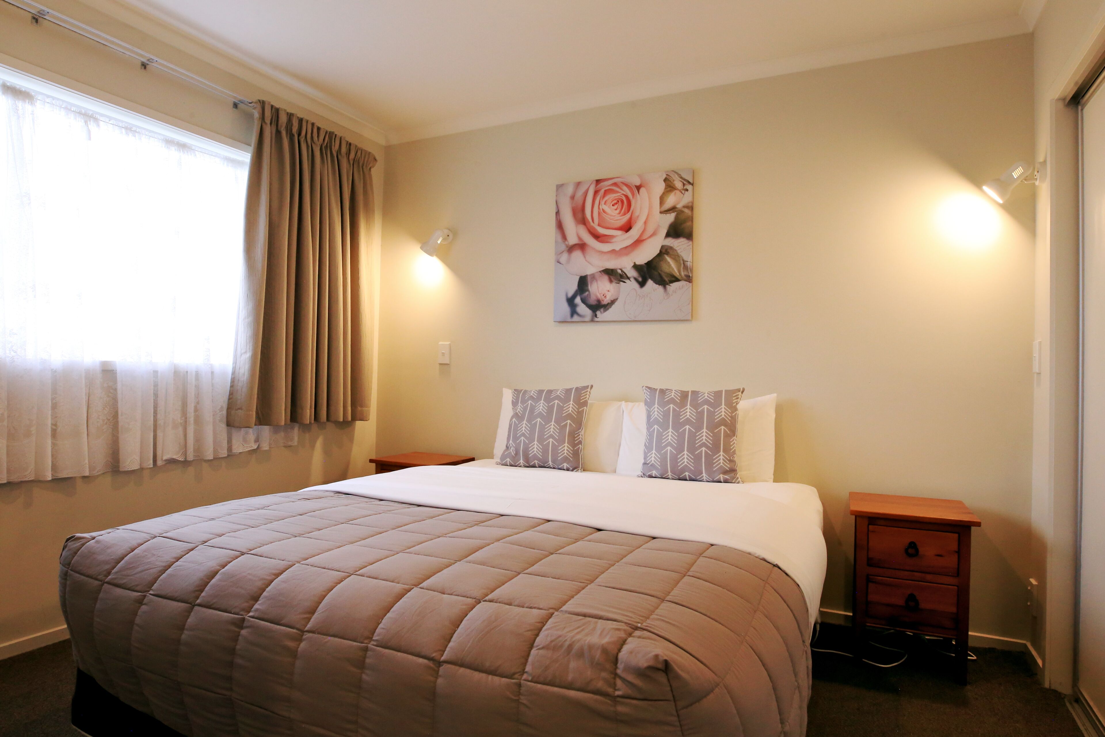 A bedroom with a bed, two nightstands, a wall-mounted light, a window with curtains, and a framed picture on the wall.