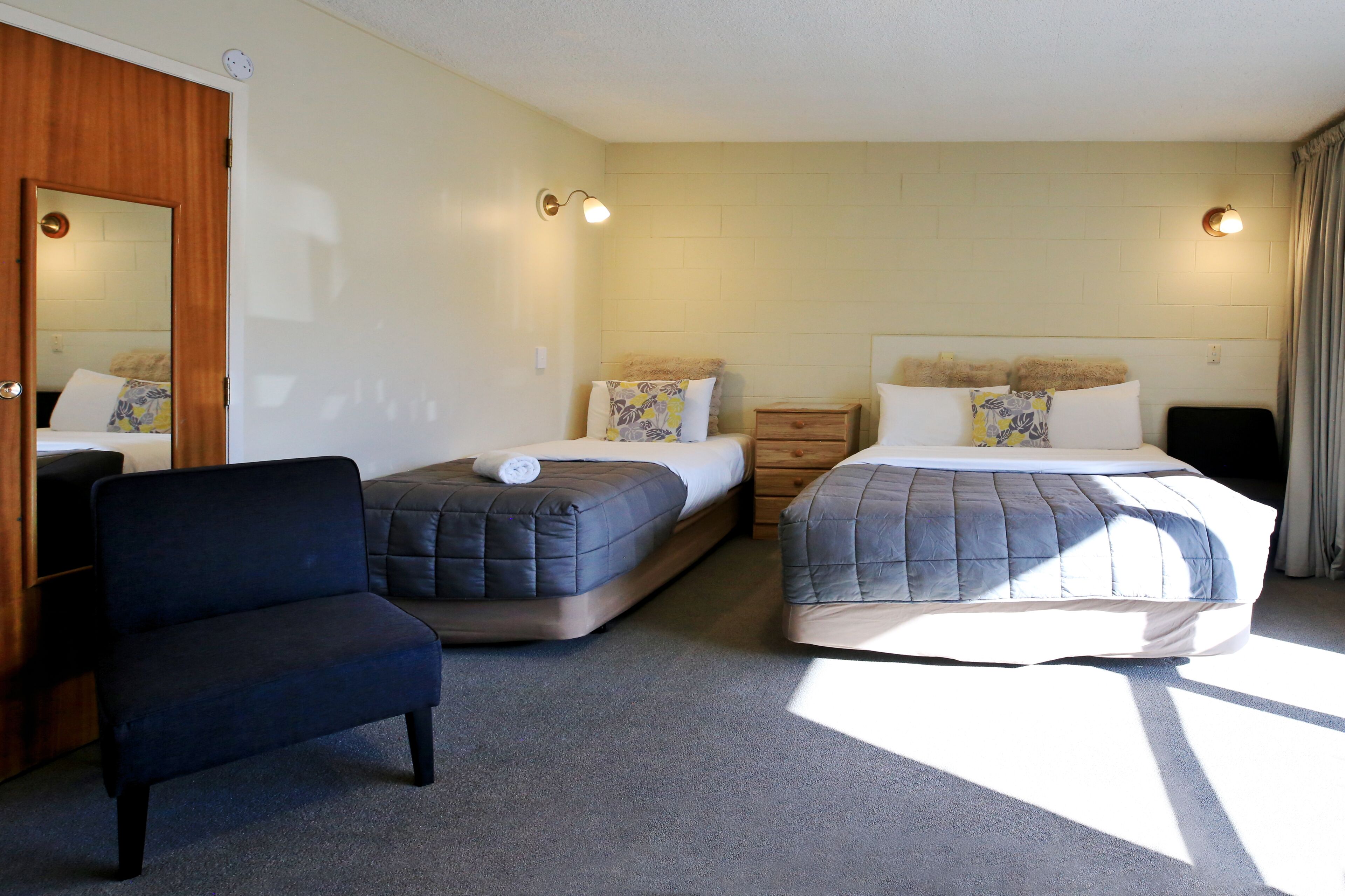 A hotel room with two beds, a chair, a wooden wardrobe, and a mirror.